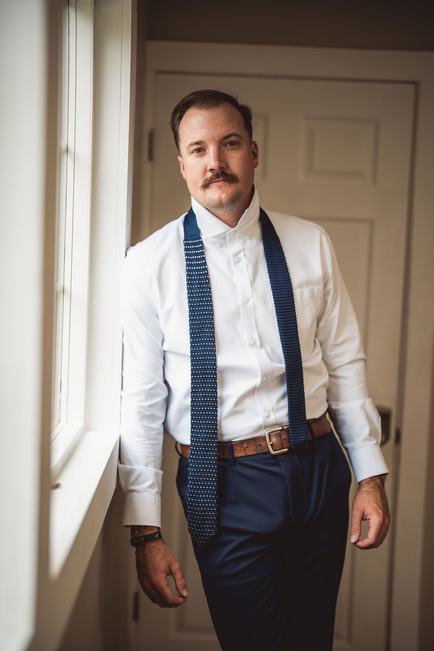 A man in formal attire adjusts his navy blue polka dot tie while getting dressed.