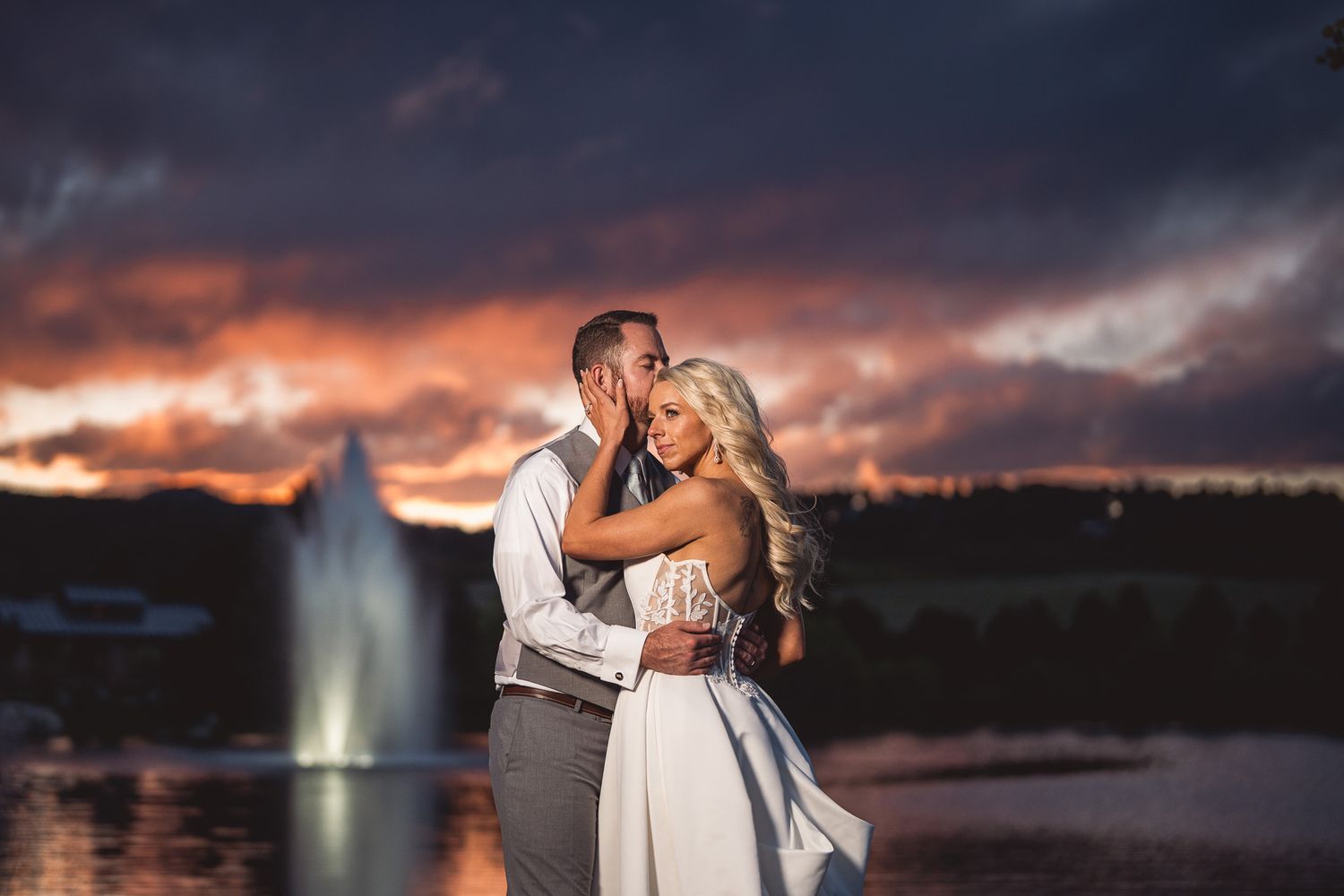 Bride and groom embrace at sunset by a fountain, with dramatic purple and orange clouds in the background.