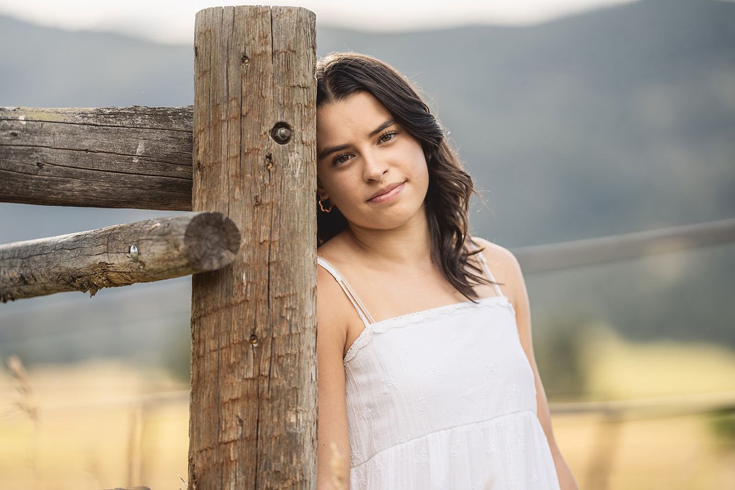Young woman in white sundress leaning against rustic wooden fence post in scenic mountain meadow.