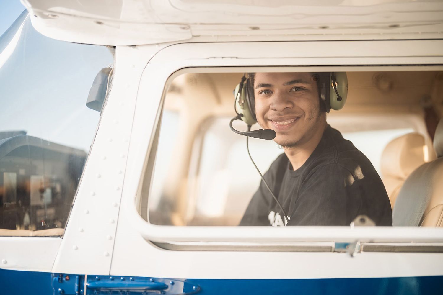 Young male pilot smiling in cockpit of small aircraft wearing headset on sunny day.