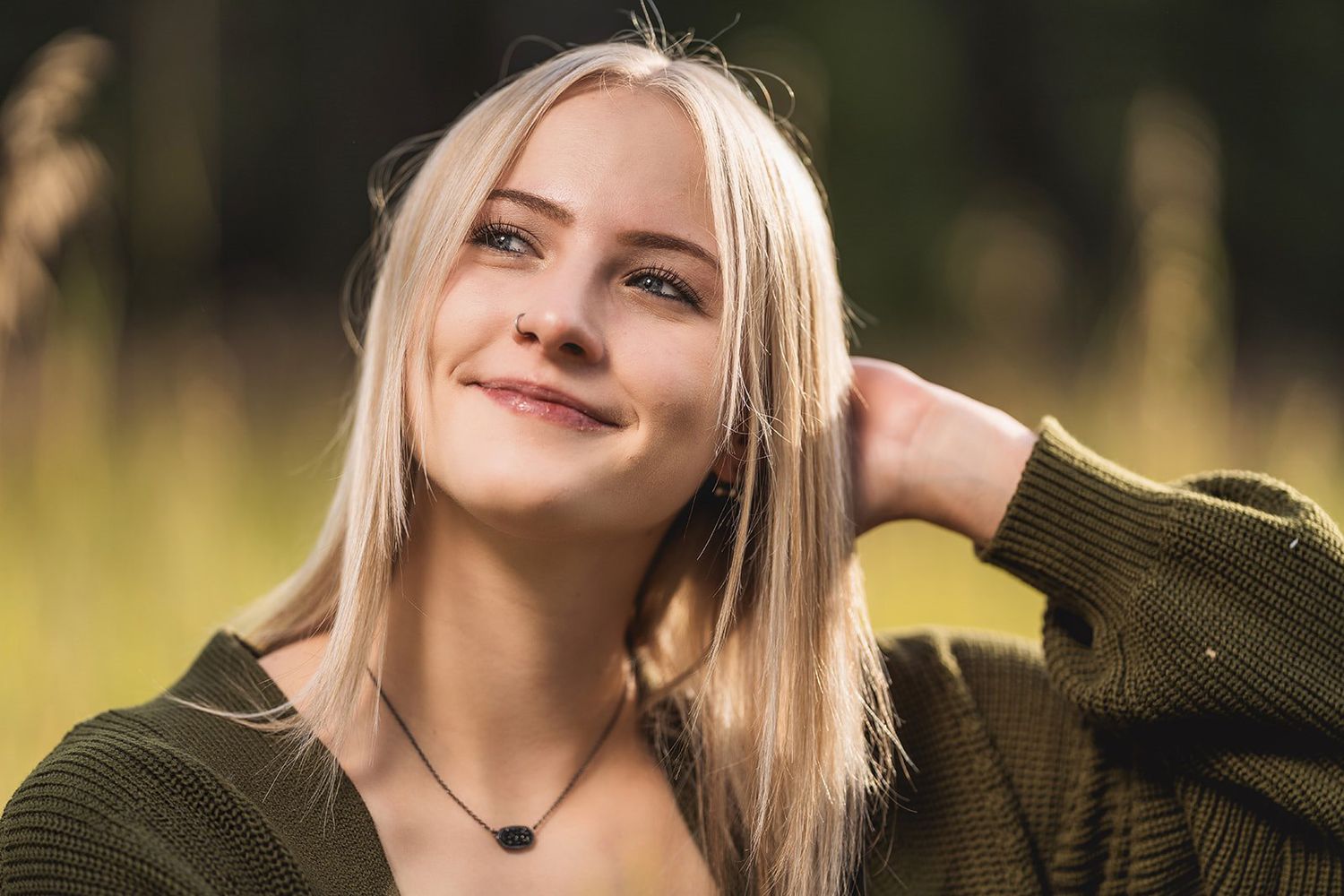 Smiling young blonde woman in olive cardigan with black pendant necklace poses outdoors in golden sunlight.