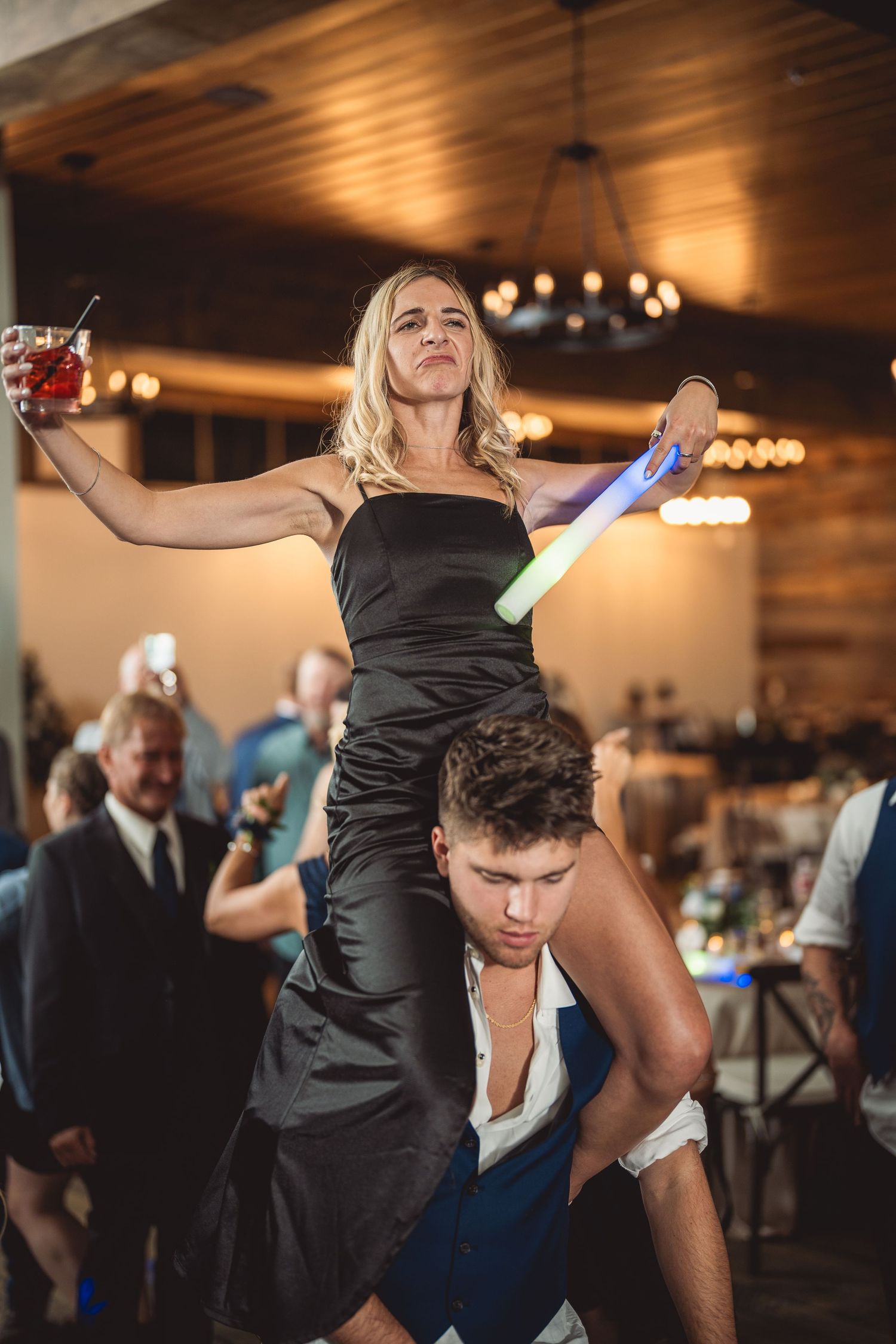 Woman in black dress sits on man's shoulders holding a cocktail and glow stick at a lively wedding reception.