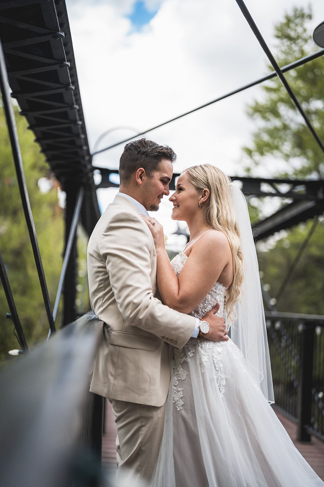 Bride and groom share a romantic moment on a bridge, with the bride in a lace gown and groom in a beige suit.