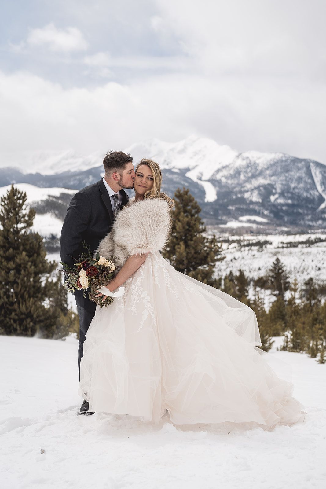 Bride and groom embrace in snowy mountain landscape, bride wearing white gown with fur wrap holding bouquet.