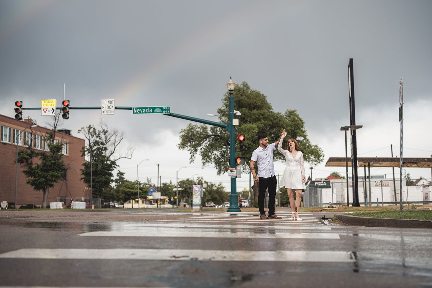 Couple walks hand-in-hand across wet intersection with rainbow in cloudy sky, romantic urban engagement photo.