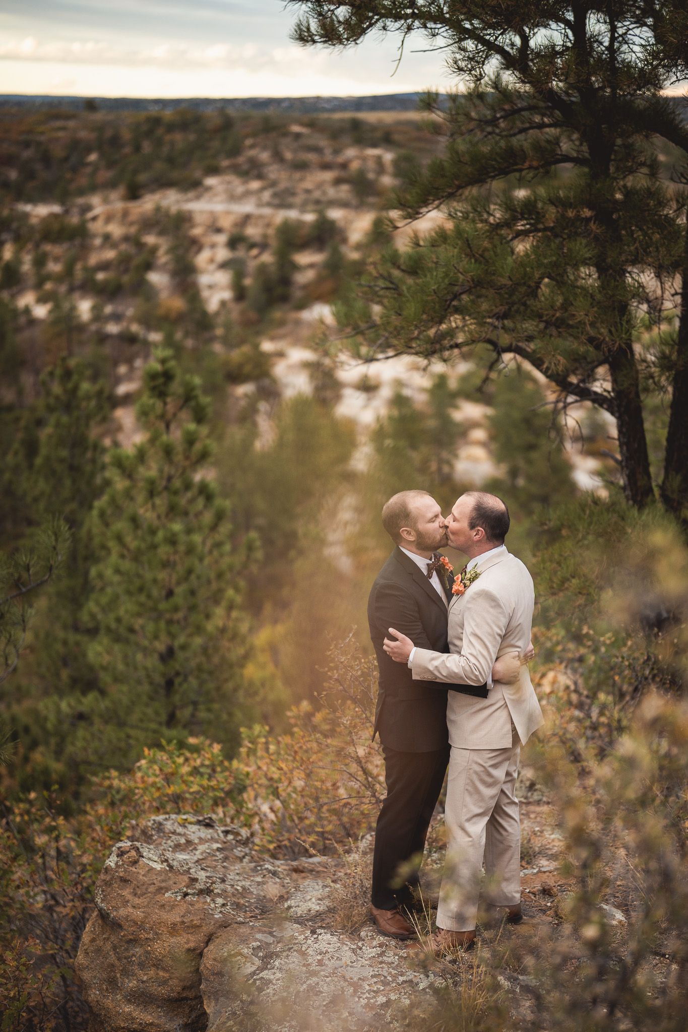 Two grooms share a kiss on a rocky overlook surrounded by pine trees and canyon views during their outdoor wedding.