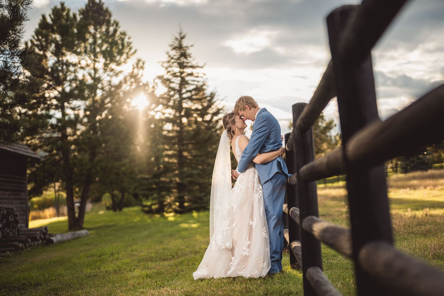 Bride and groom share a romantic kiss at sunset near a rustic fence surrounded by tall pine trees on a lush green lawn.