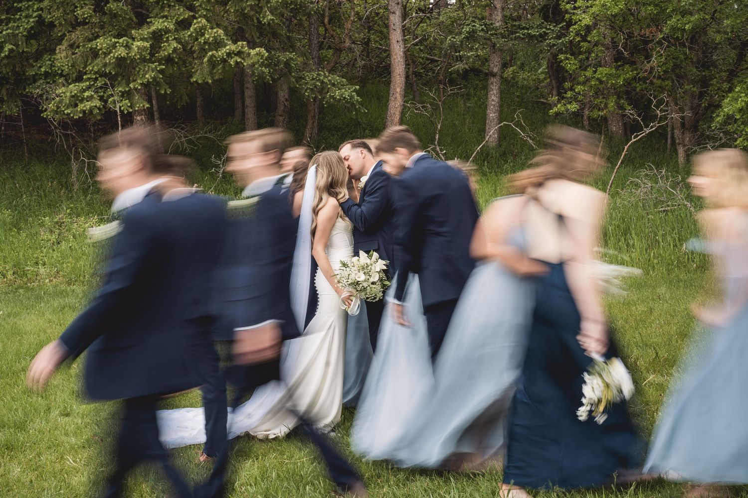 Bride and groom share a kiss while wedding party walks past in motion blur on green grass.