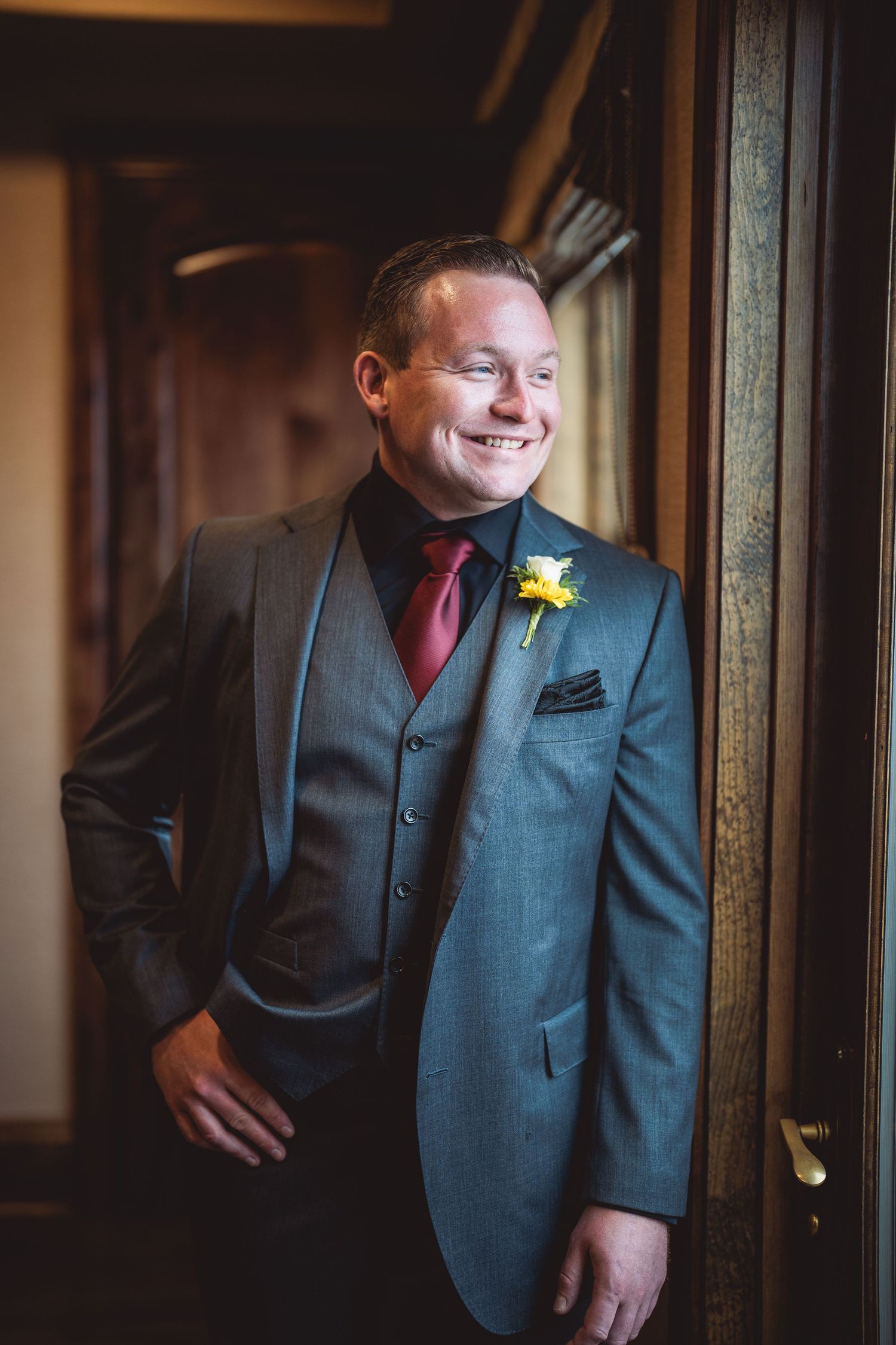 A groom in a grey suit and red tie smiling while leaning against a window.