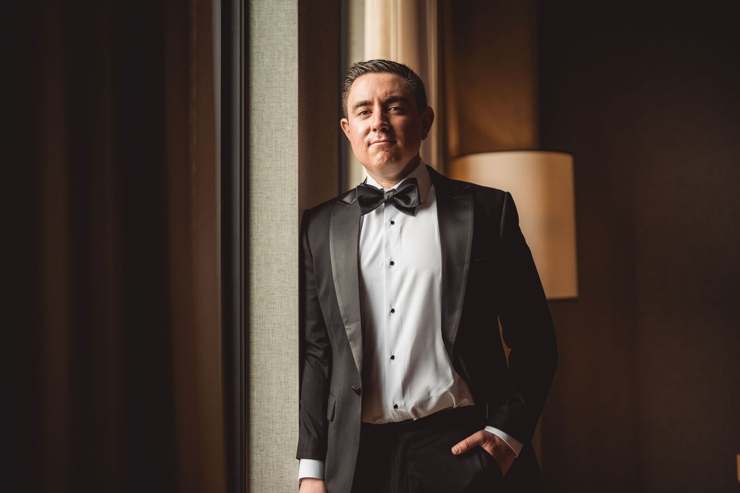 Person in formal attire smiling while posing near window with warm lighting.