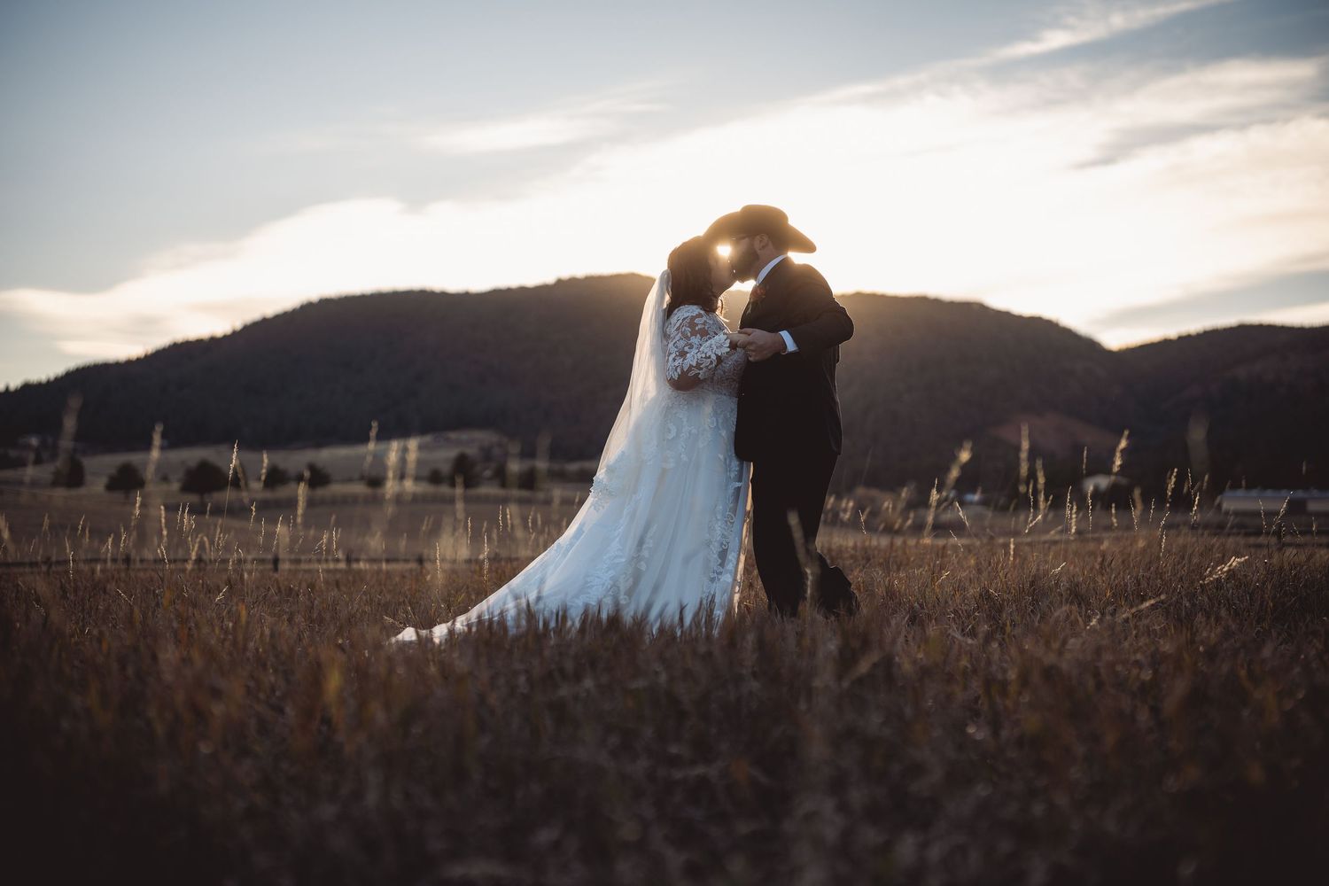 Silhouetted couple embracing in golden field at sunset with mountains.