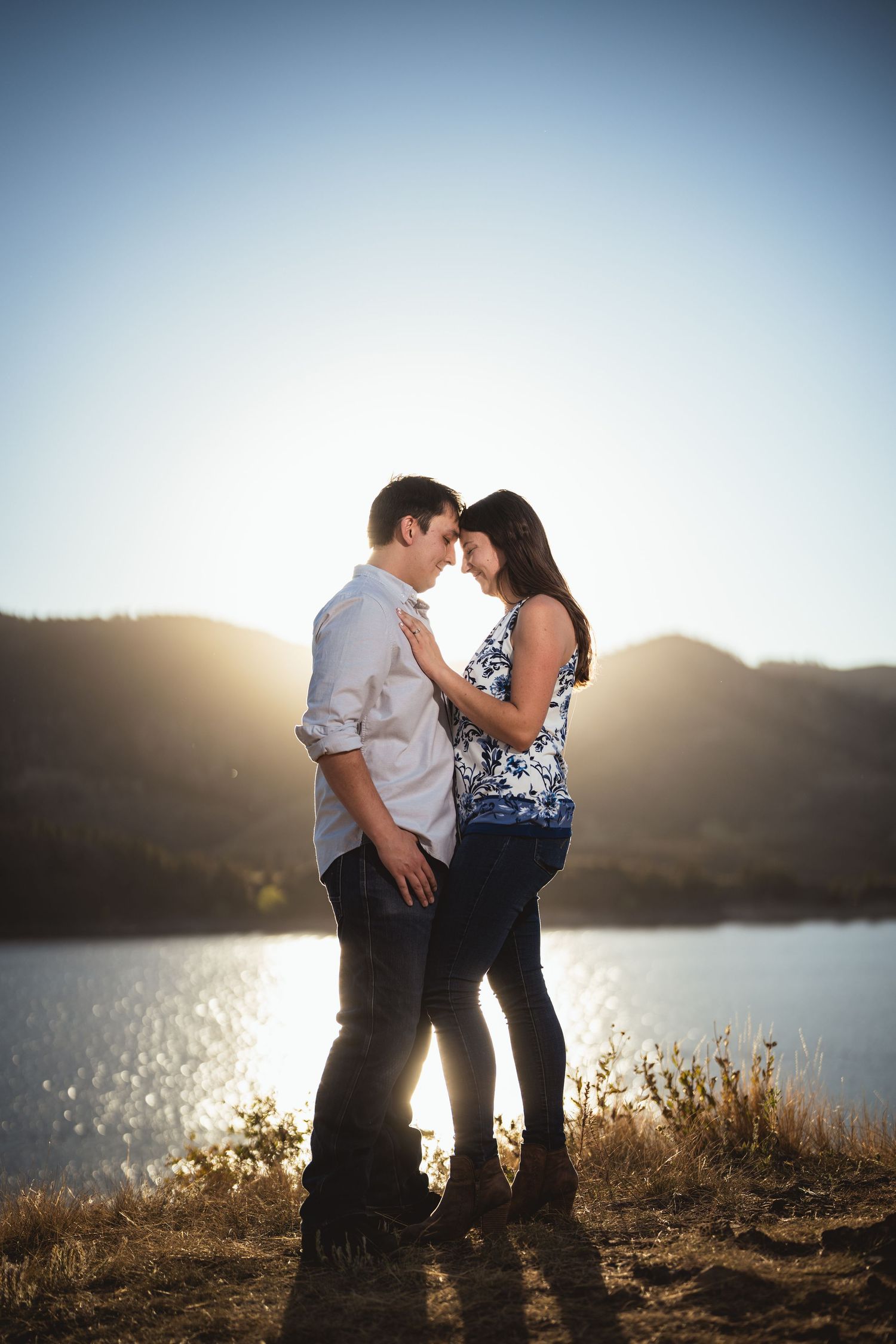 Couple embraces on a hillside overlooking a lake during golden hour sunset.