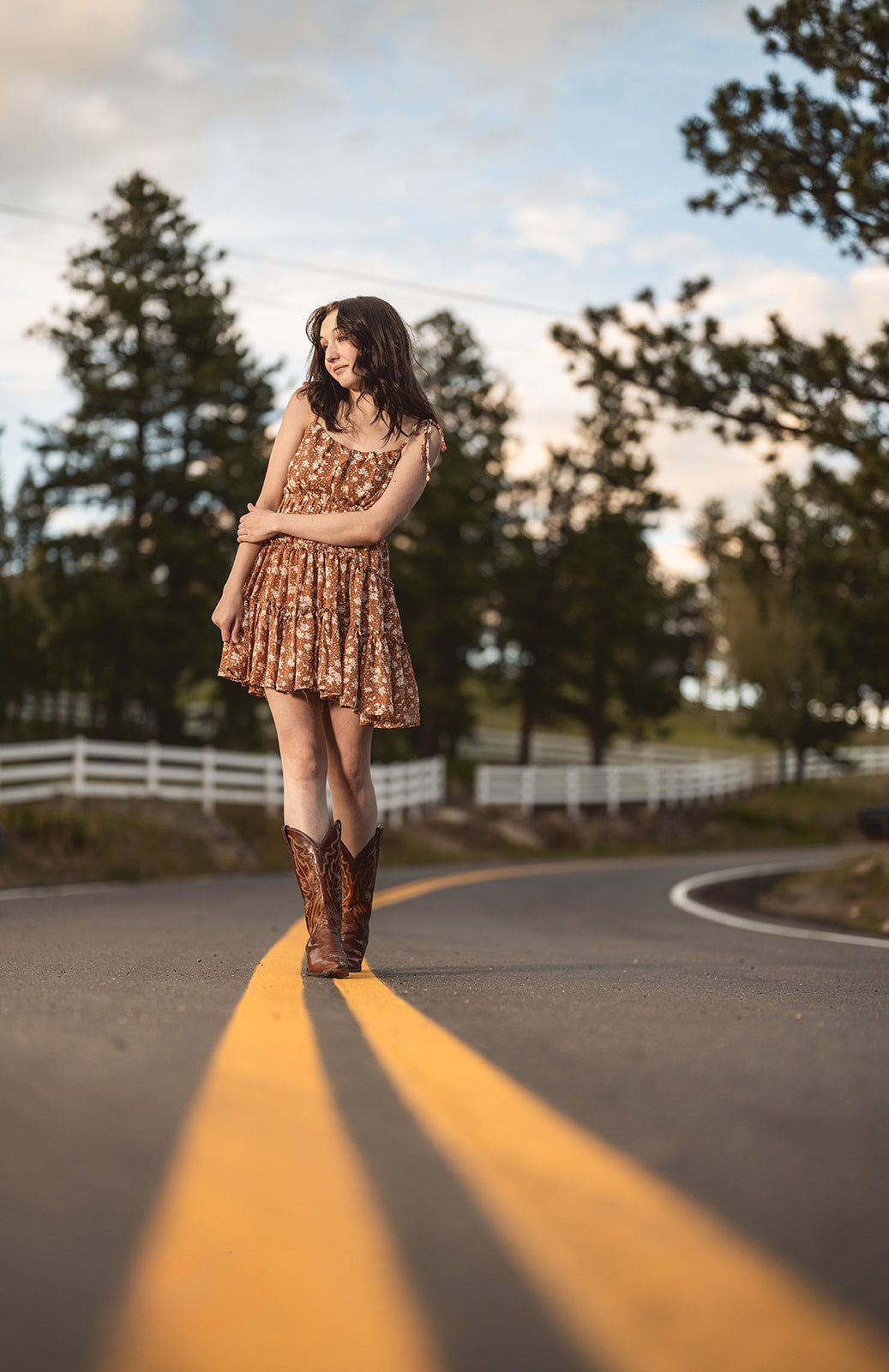Brunette woman in floral dress and cowboy boots walking on road at golden hour.
