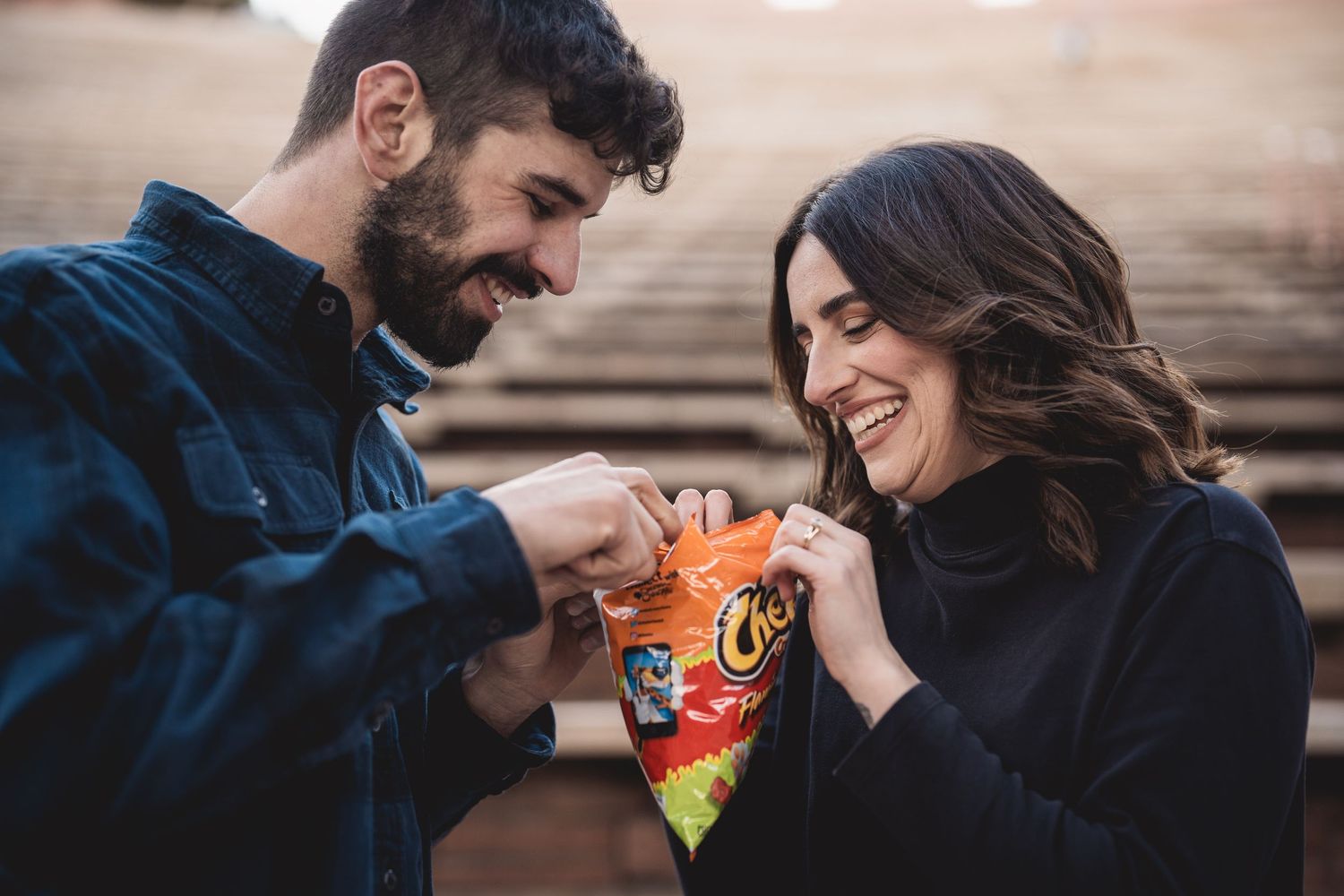 A smiling couple laughs while opening a bag of Cheetos together outdoors on steps.