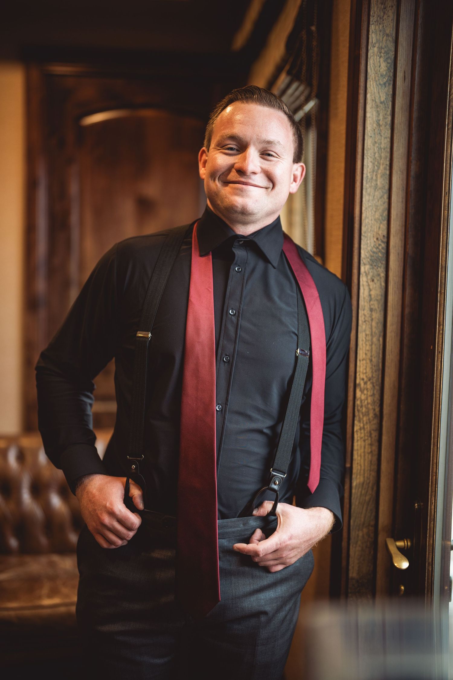 Smiling groom in black shirt, burgundy tie, and suspenders posing confidently near a wooden door in elegant venue.