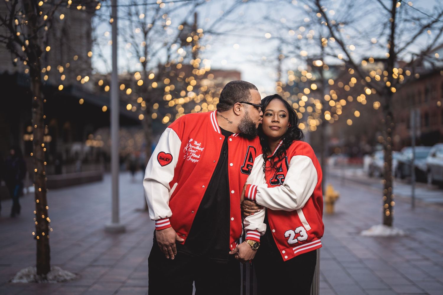 Couple in matching red varsity jackets sharing a kiss outdoors with bokeh string lights in background.