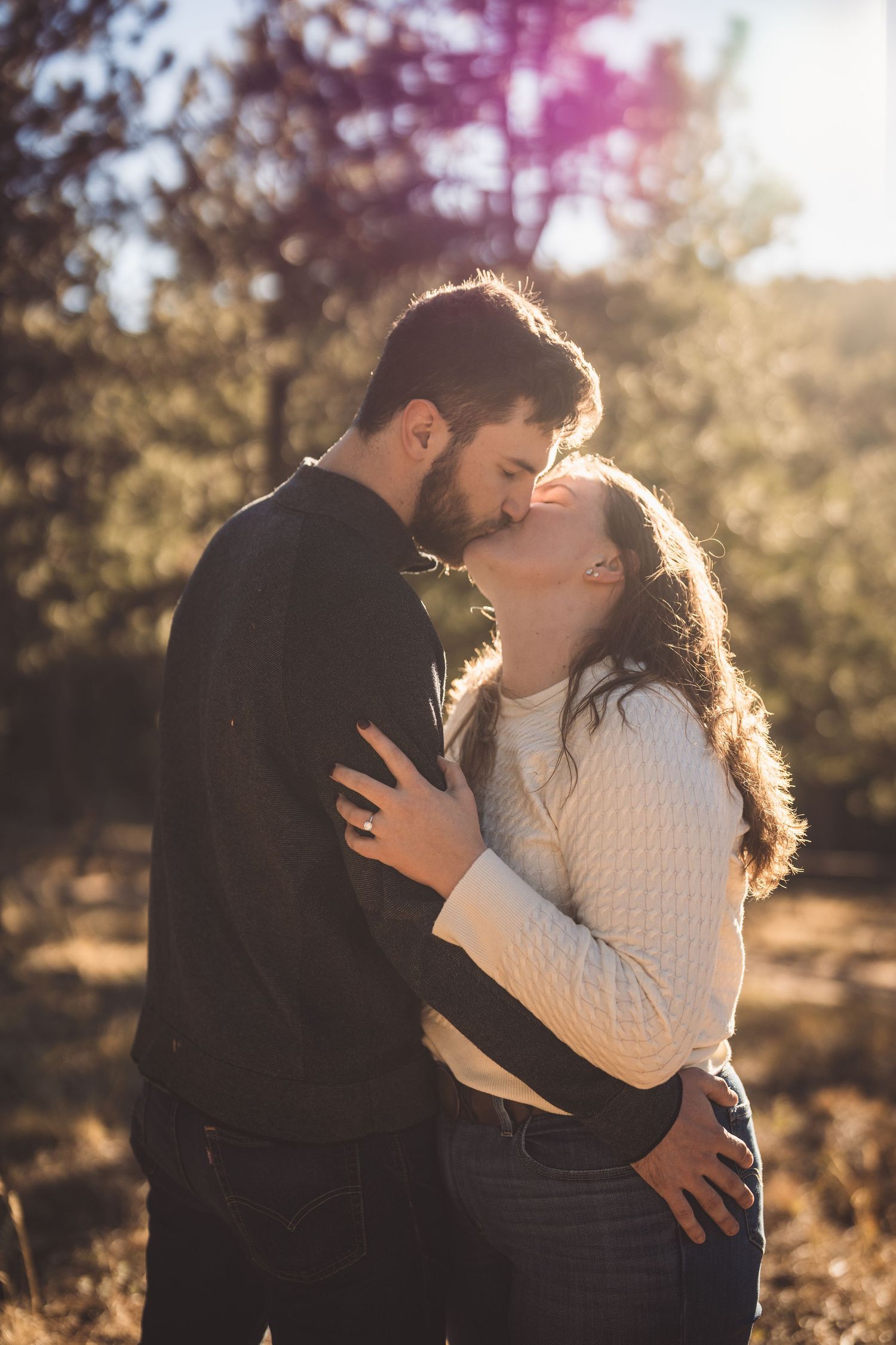 A couple shares a romantic kiss outdoors in golden sunlight, surrounded by trees in a warm autumn setting.