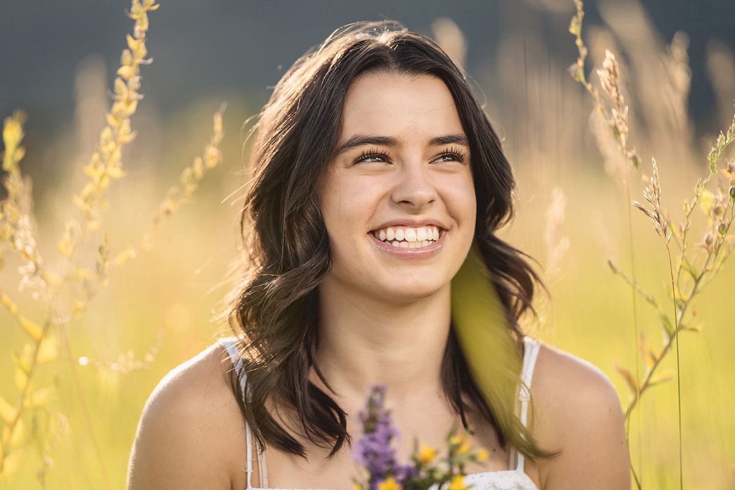 Smiling young woman with dark hair holding wildflowers in a sunlit golden field.