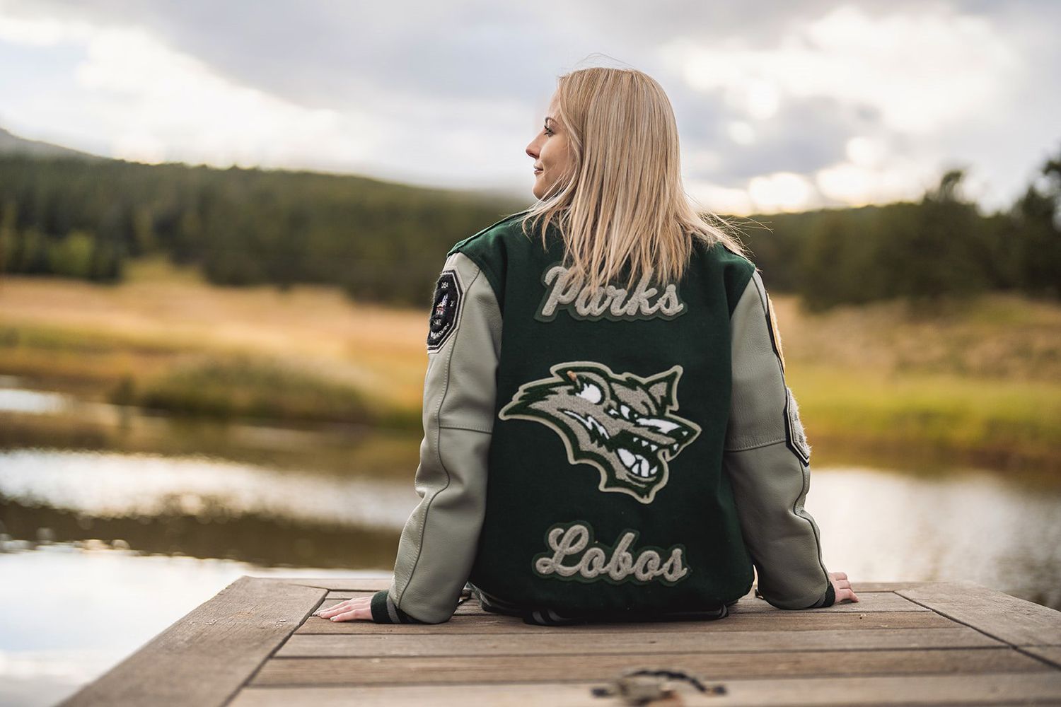 Blonde girl sitting on a dock wearing a green Parks Lobos varsity jacket near a mountain lake.