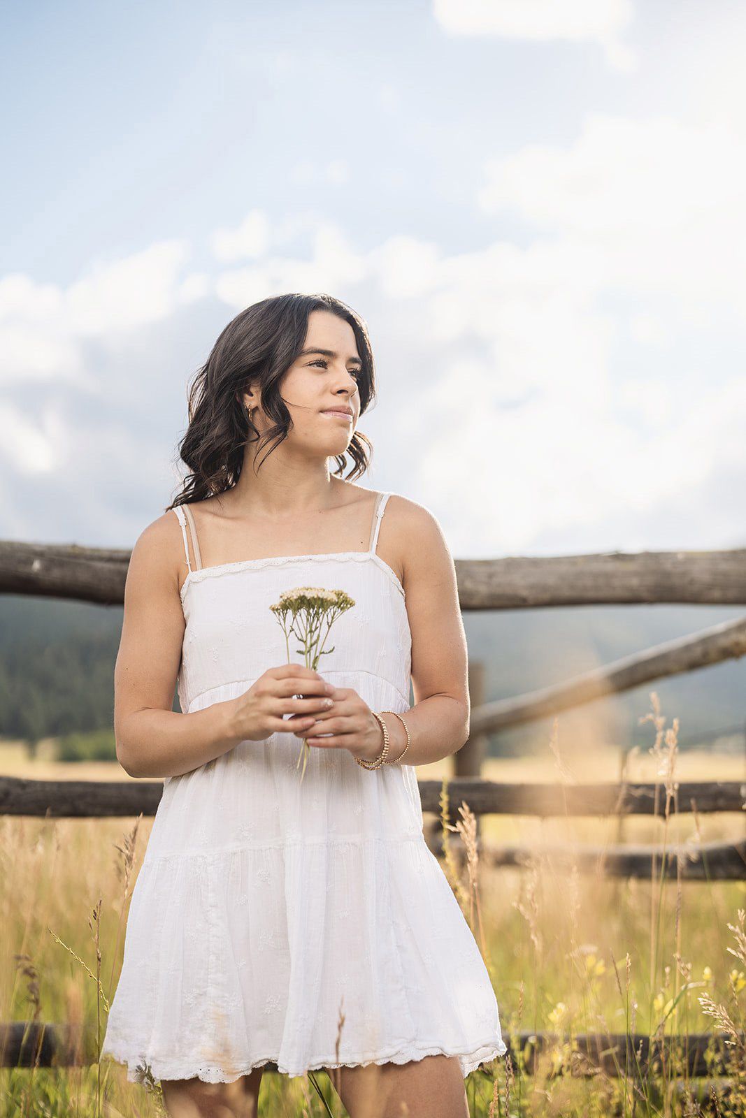 Young woman in white sundress holding wildflowers in mountain meadow with wooden fence and blue sky.