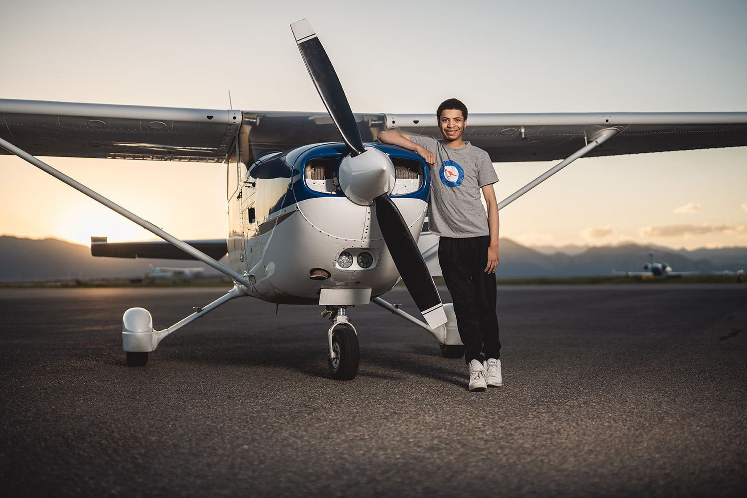 Young man standing proudly in front of a small propeller aircraft on a runway at sunset.