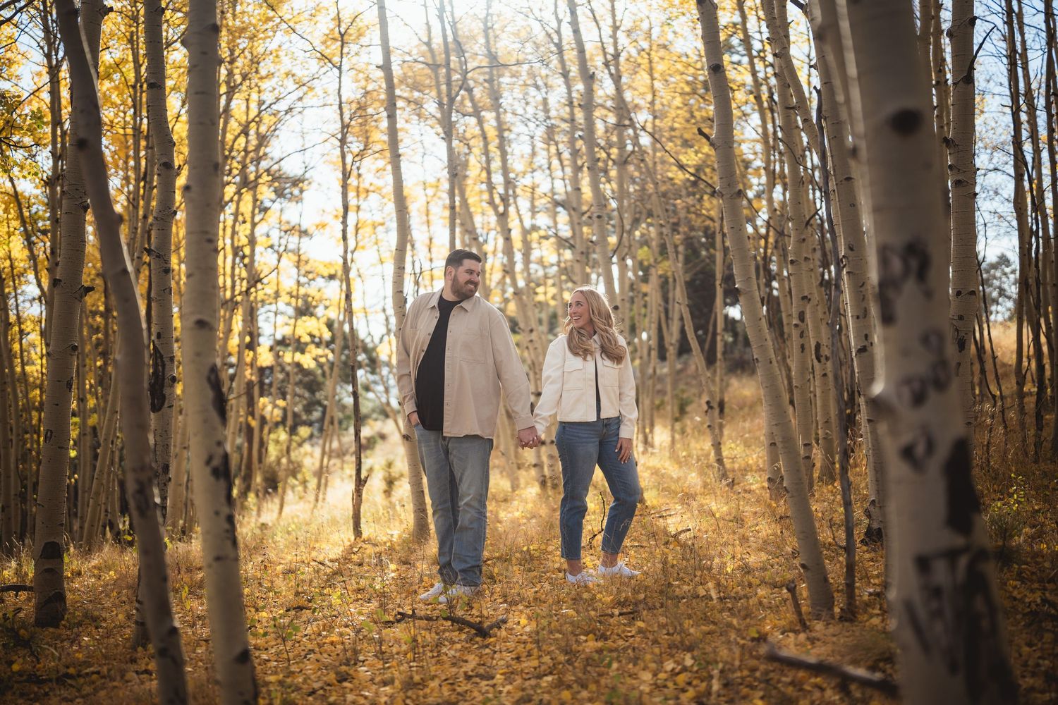 Couple holding hands walking through golden aspen forest in autumn, surrounded by yellow leaves and white birch trees.