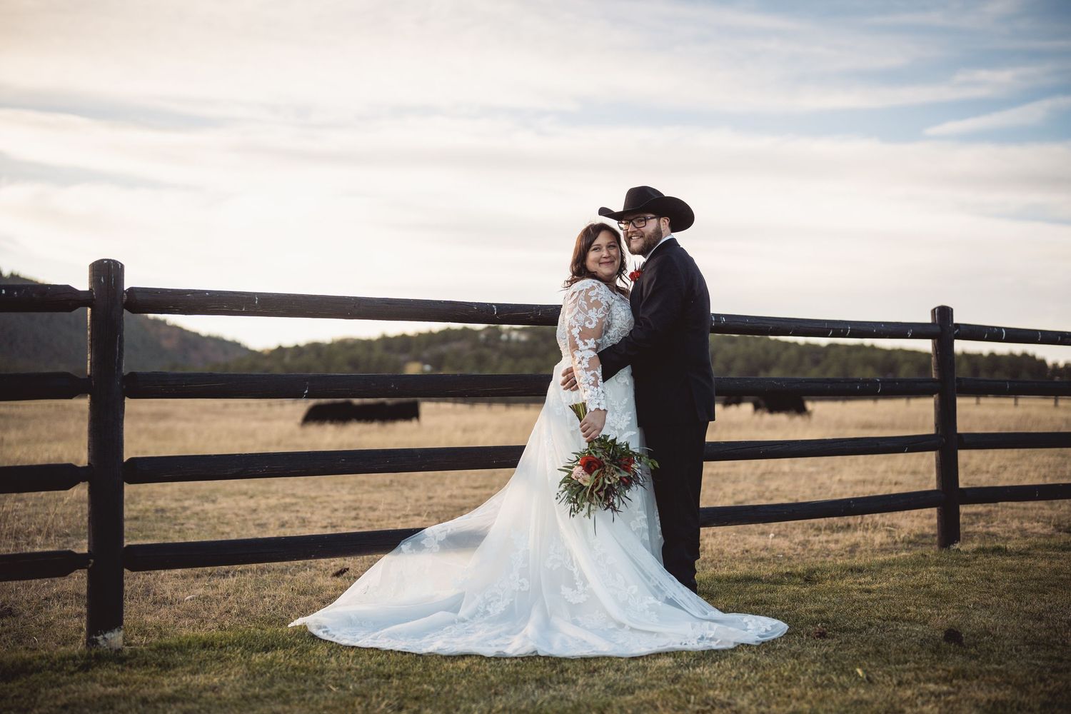 Bride and groom pose by rustic wooden fence on a farm, bride in lace gown holding bouquet, groom in black cowboy hat.