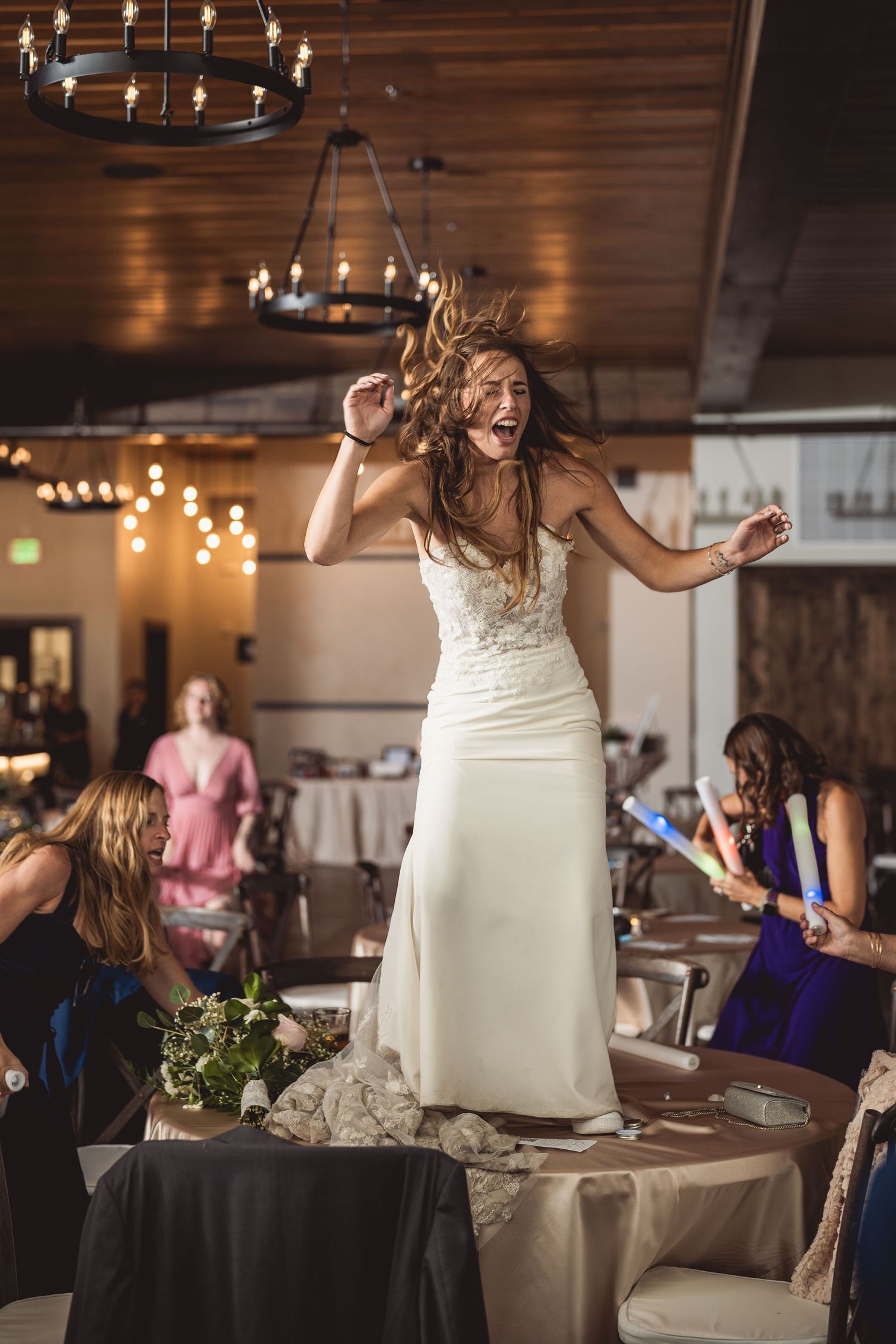 Joyful bride in white gown dancing energetically at wedding reception with guests and warm string lights in background.