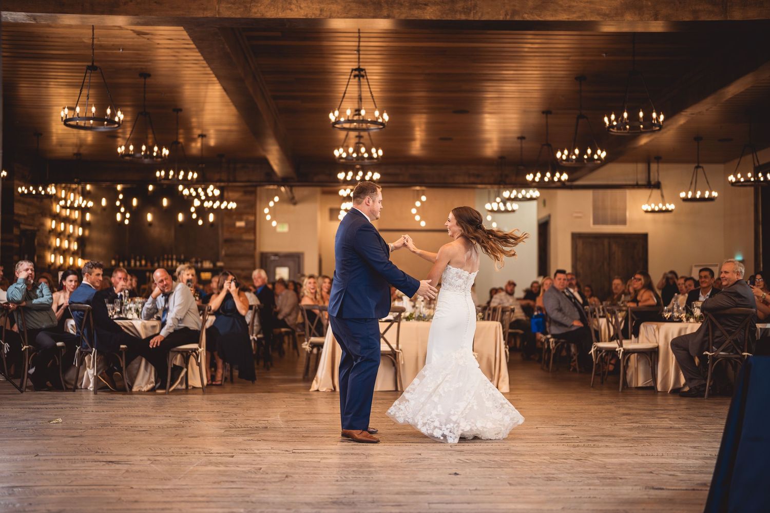 Bride and groom share their first dance in an elegant rustic reception hall with chandeliers and seated wedding guests.