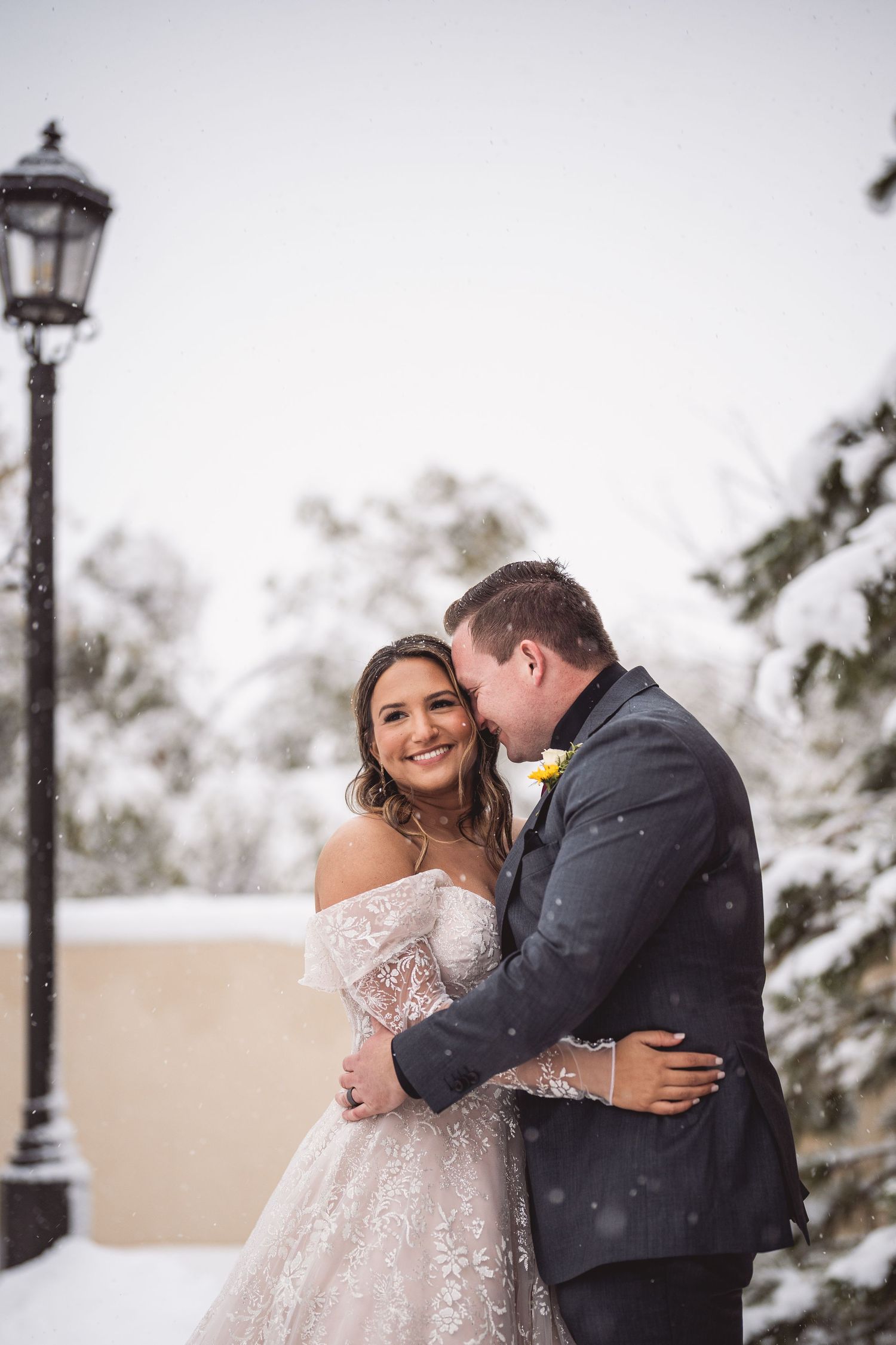 Groom kisses bride on cheek in a romantic snowy winter wedding portrait with lamp post.