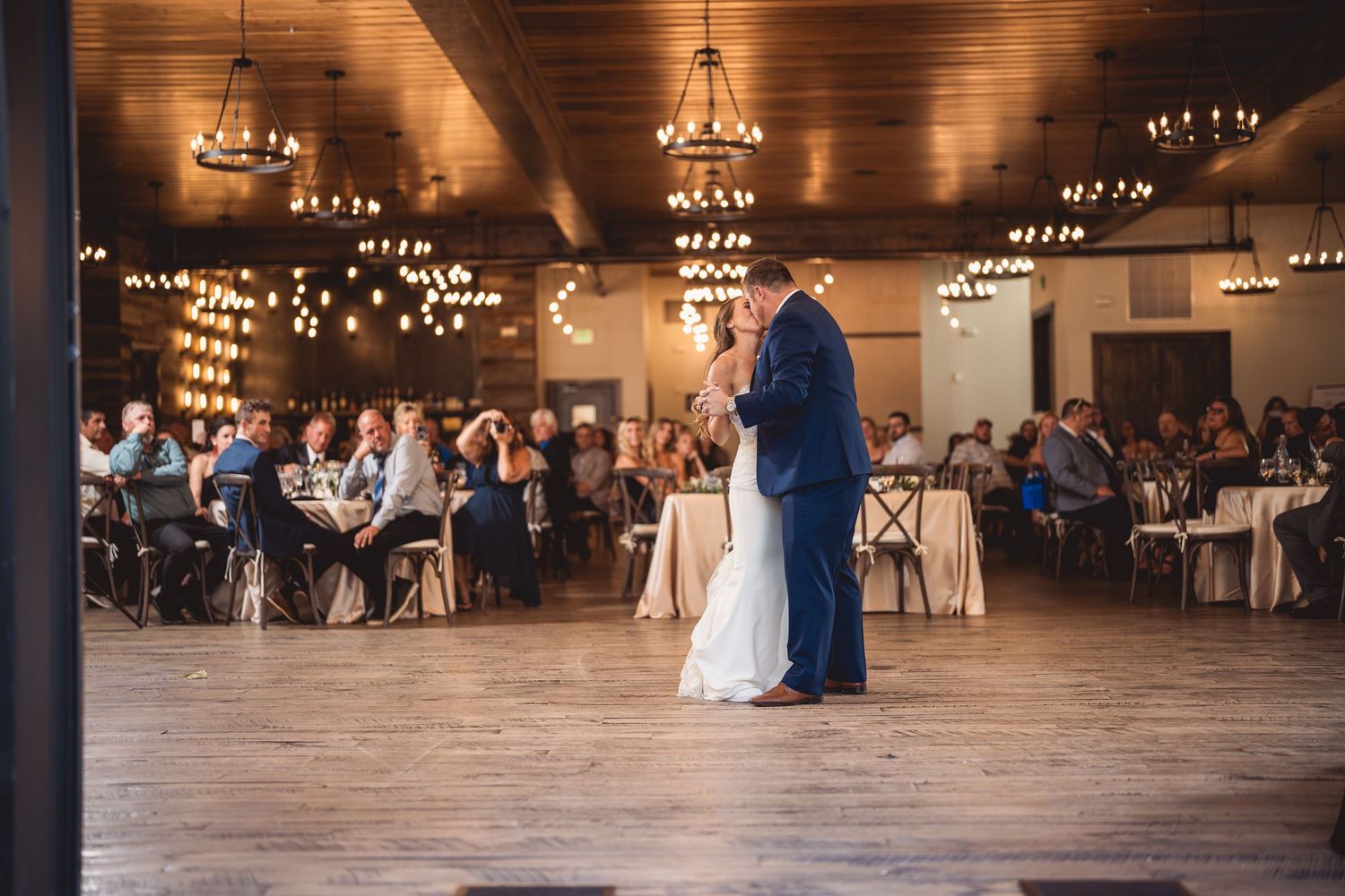 Couple shares first dance in elegant rustic barn wedding venue with chandeliers and seated guests.