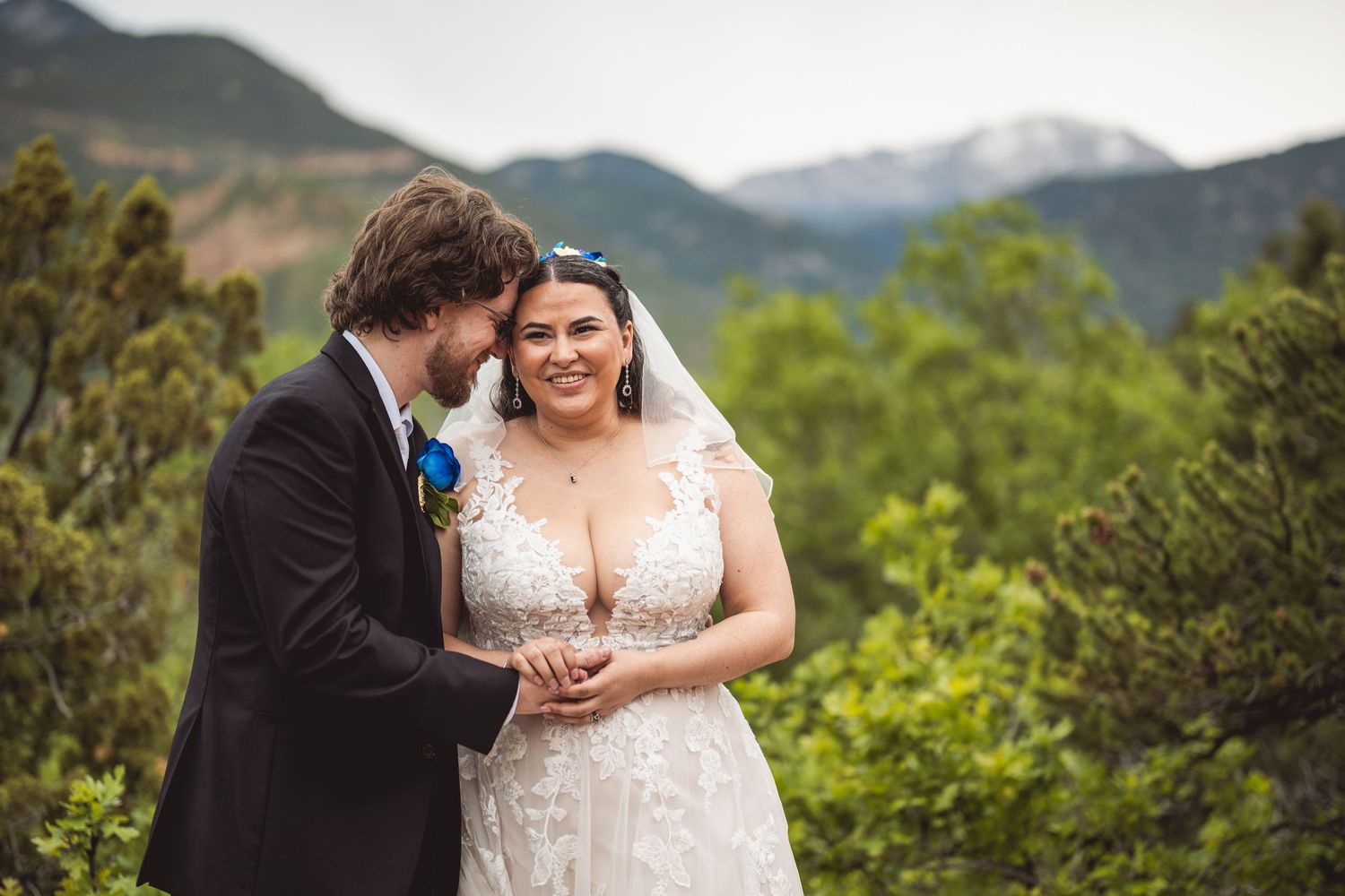 Smiling bride in lace gown with groom holding hands before a scenic mountain backdrop at outdoor wedding.