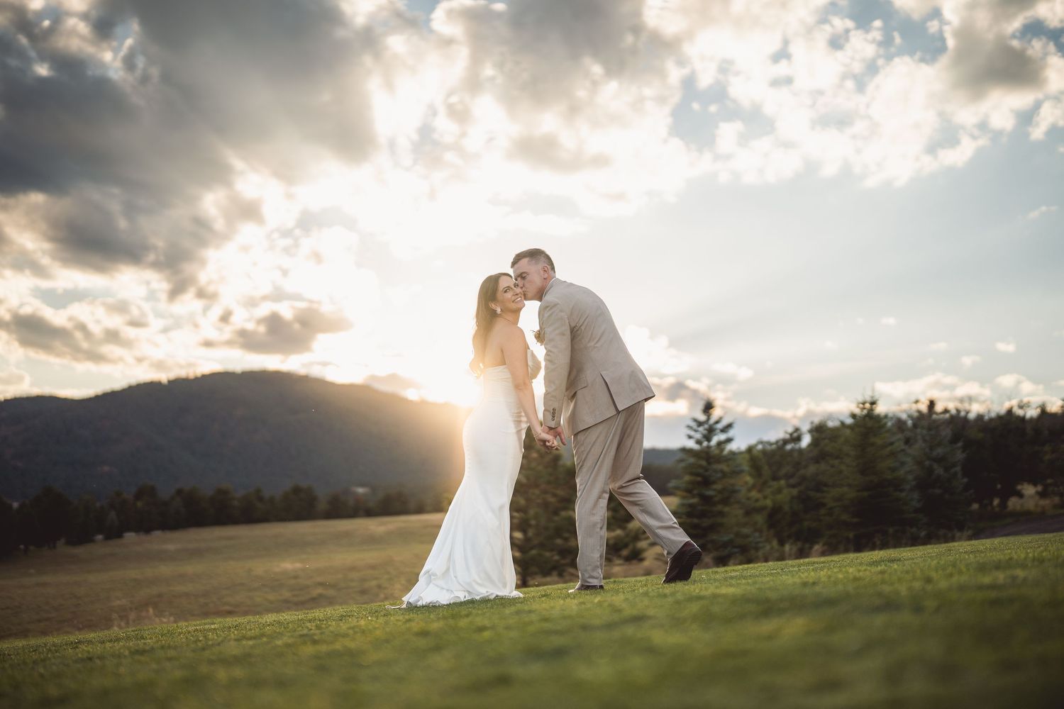 Bride and groom share a romantic kiss at sunset on a lush green field with mountain views.