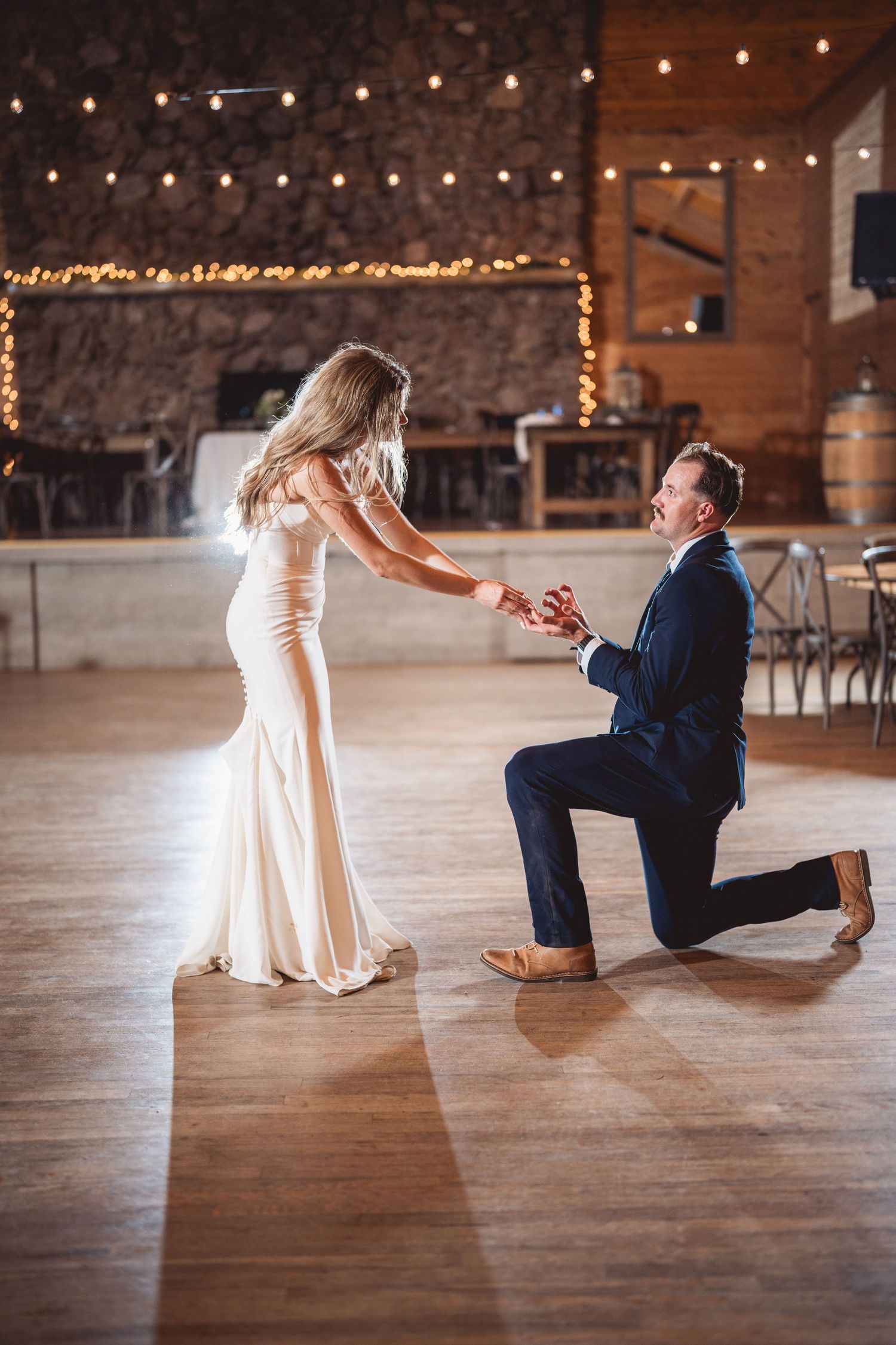 Romantic proposal moment captured on rustic wooden floor under string lights in historic barn venue.