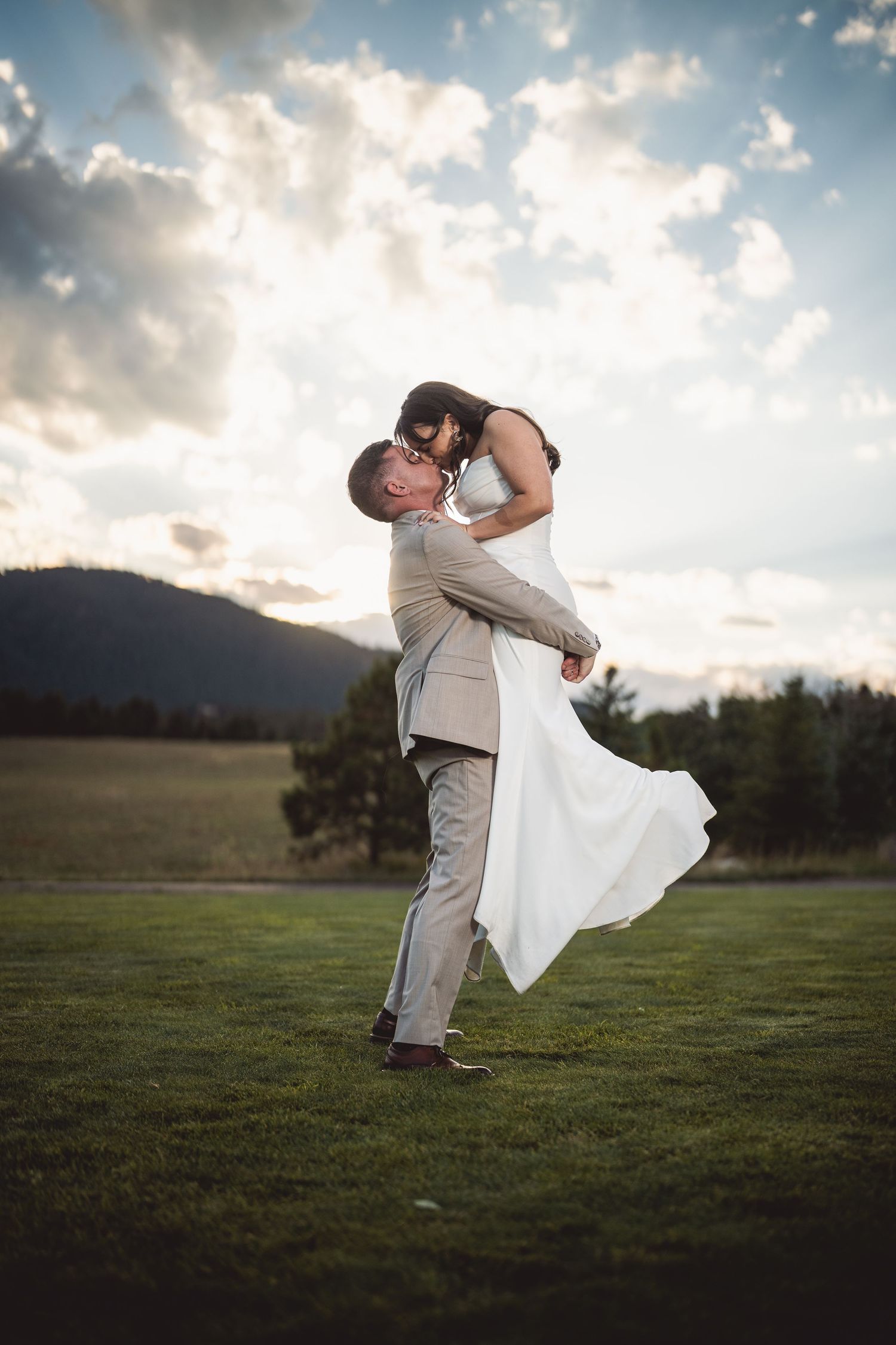 A romantic wedding couple embraces in a field at sunset with dramatic clouds in the background.