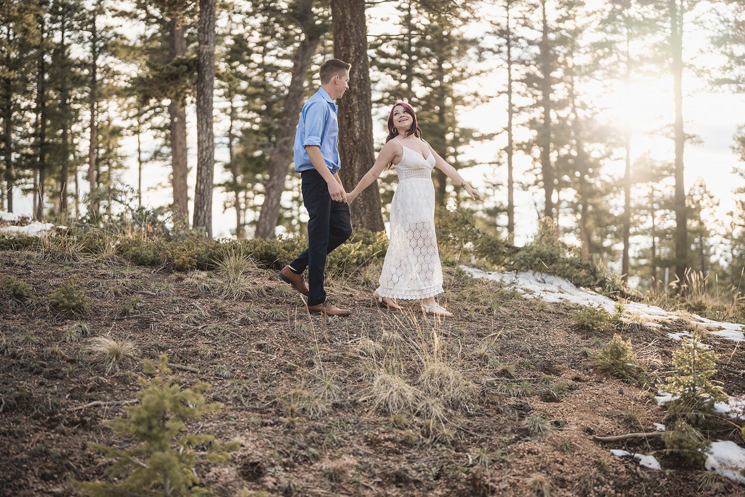 Two people walking through forest path at sunset with sunlight filtering through pine trees.