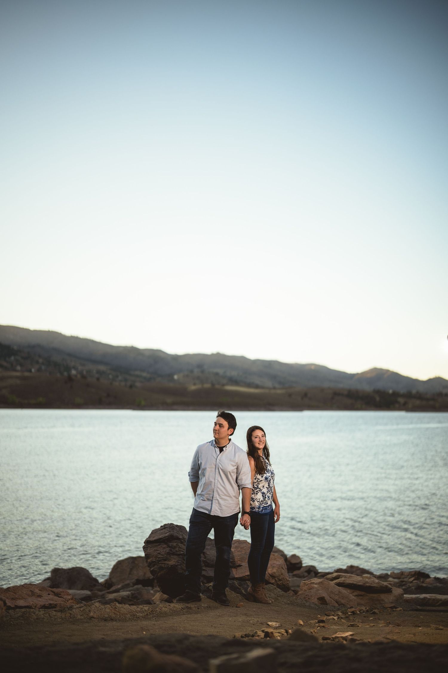 A couple poses together on rocky coastal cliffs overlooking a serene lake at sunset.
