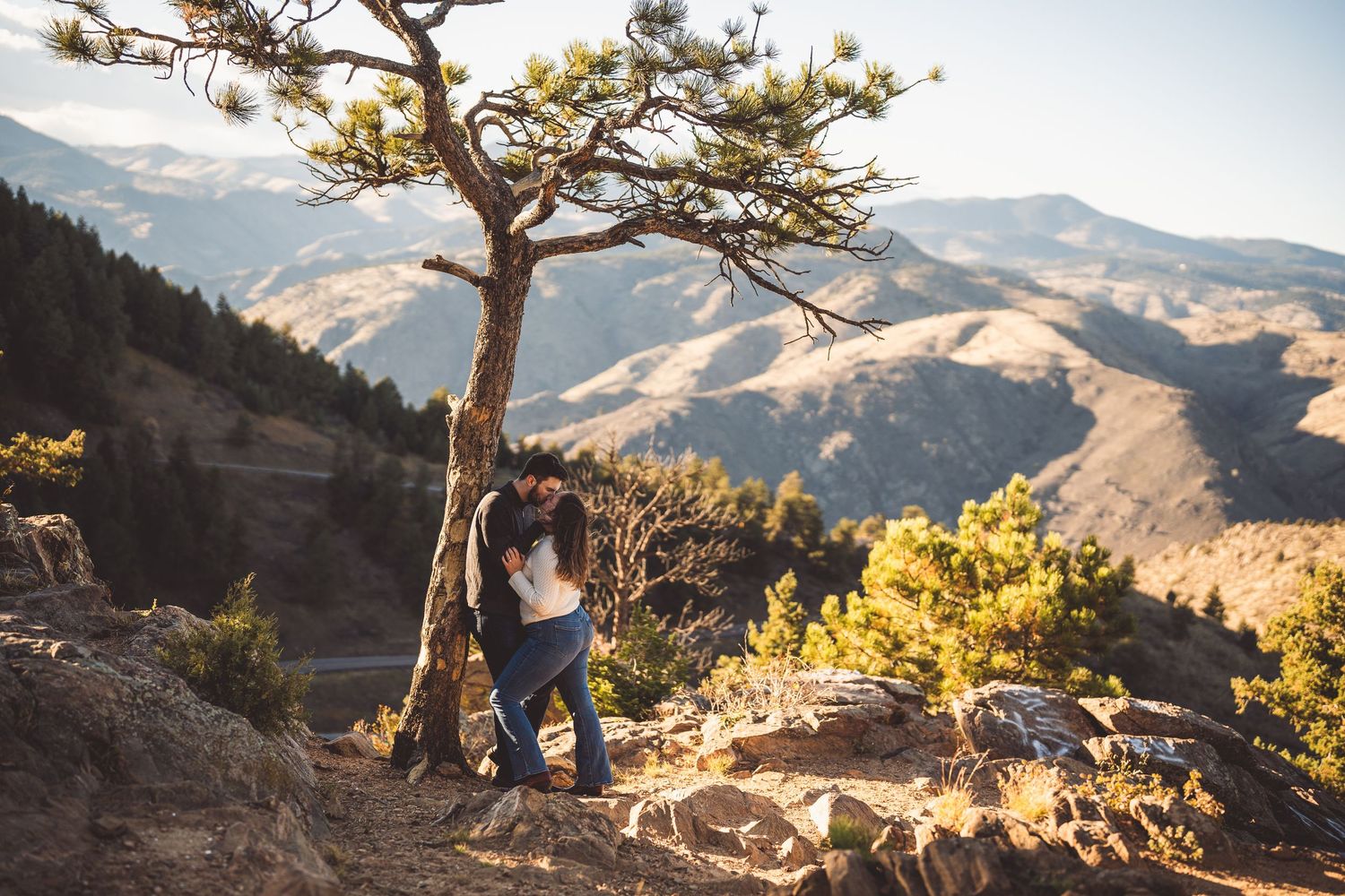 A couple shares an intimate moment under a gnarled tree with scenic mountain views at sunset.