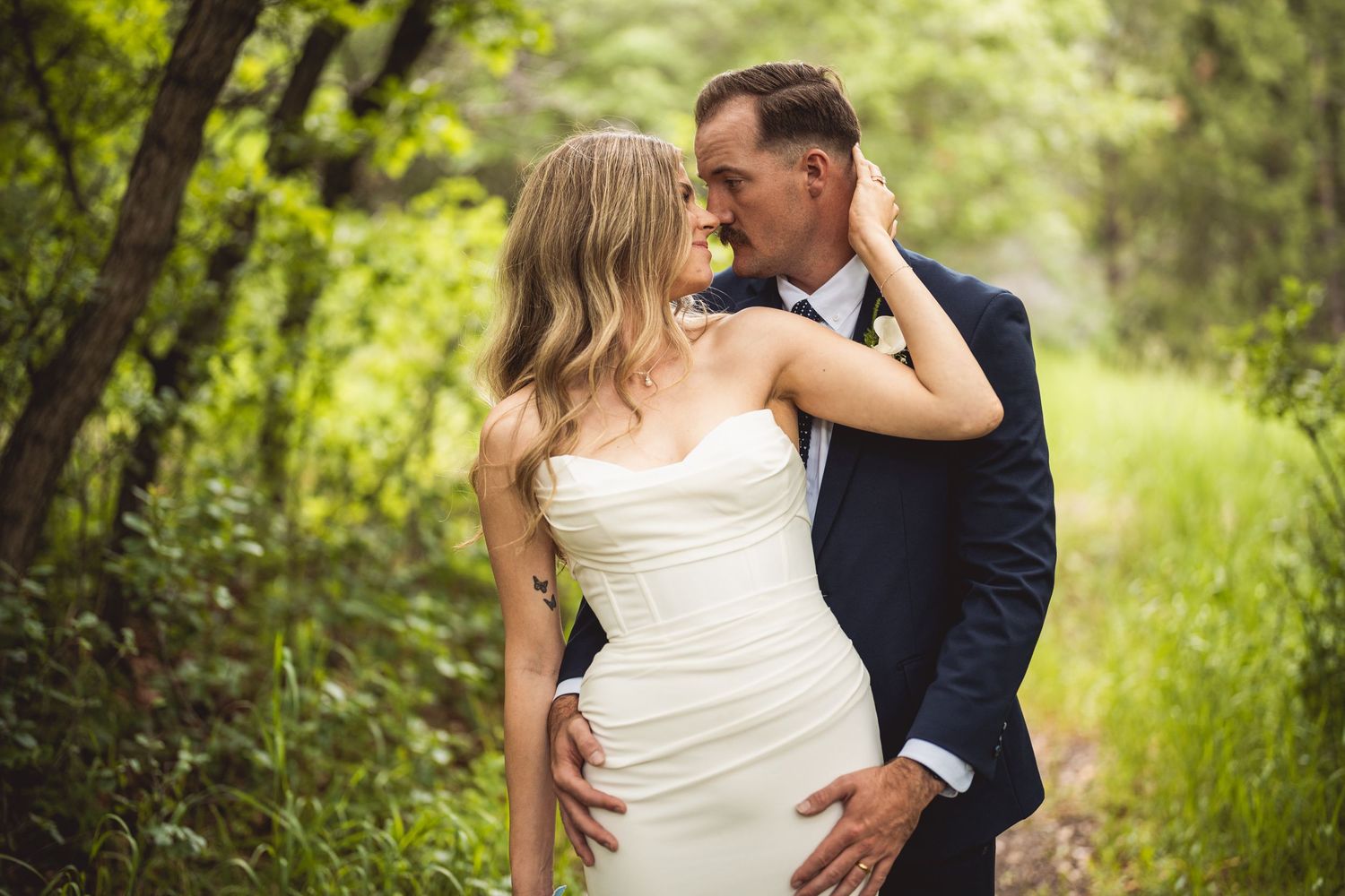 Newlyweds share an intimate moment in a lush green outdoor setting wearing classic wedding attire.