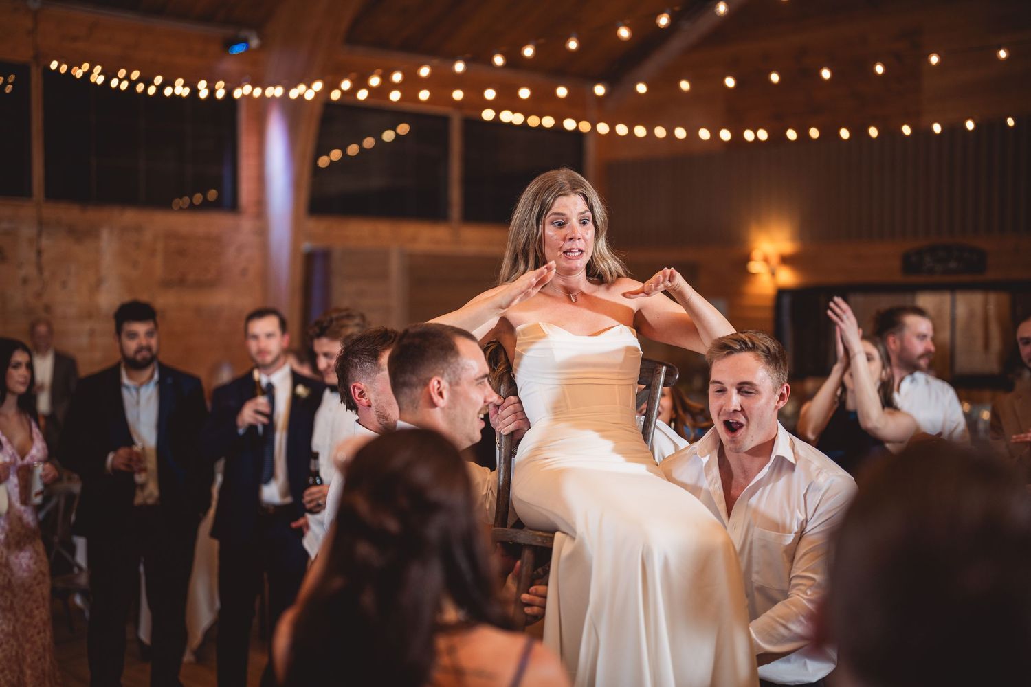 Wedding party dancing enthusiastically under string lights in rustic indoor reception space.
