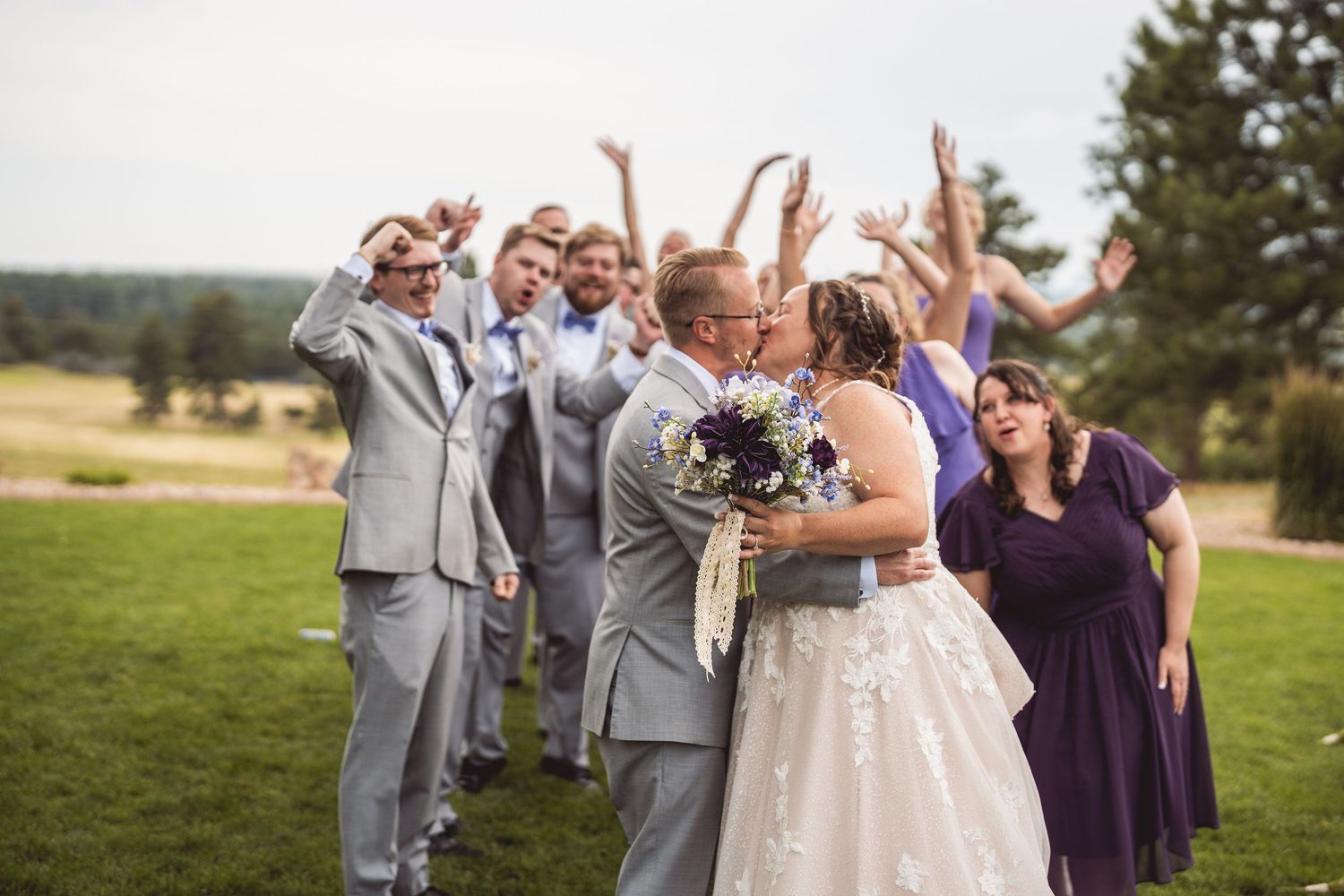 Wedding party celebrates outdoors with joyful embraces in a scenic mountain setting.