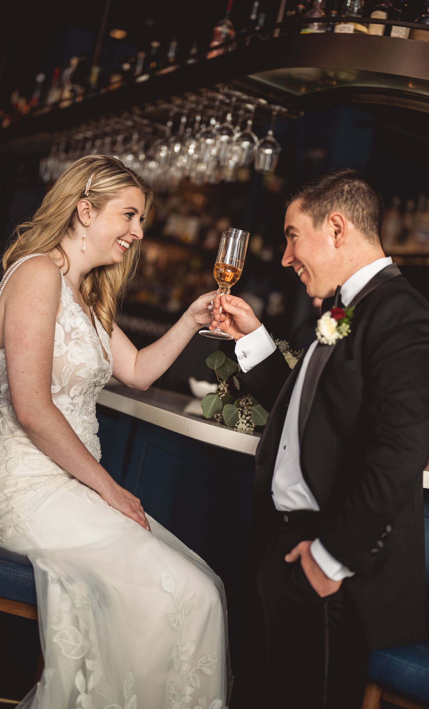 Romantic couple sharing drinks at a dimly lit bar in wedding attire.