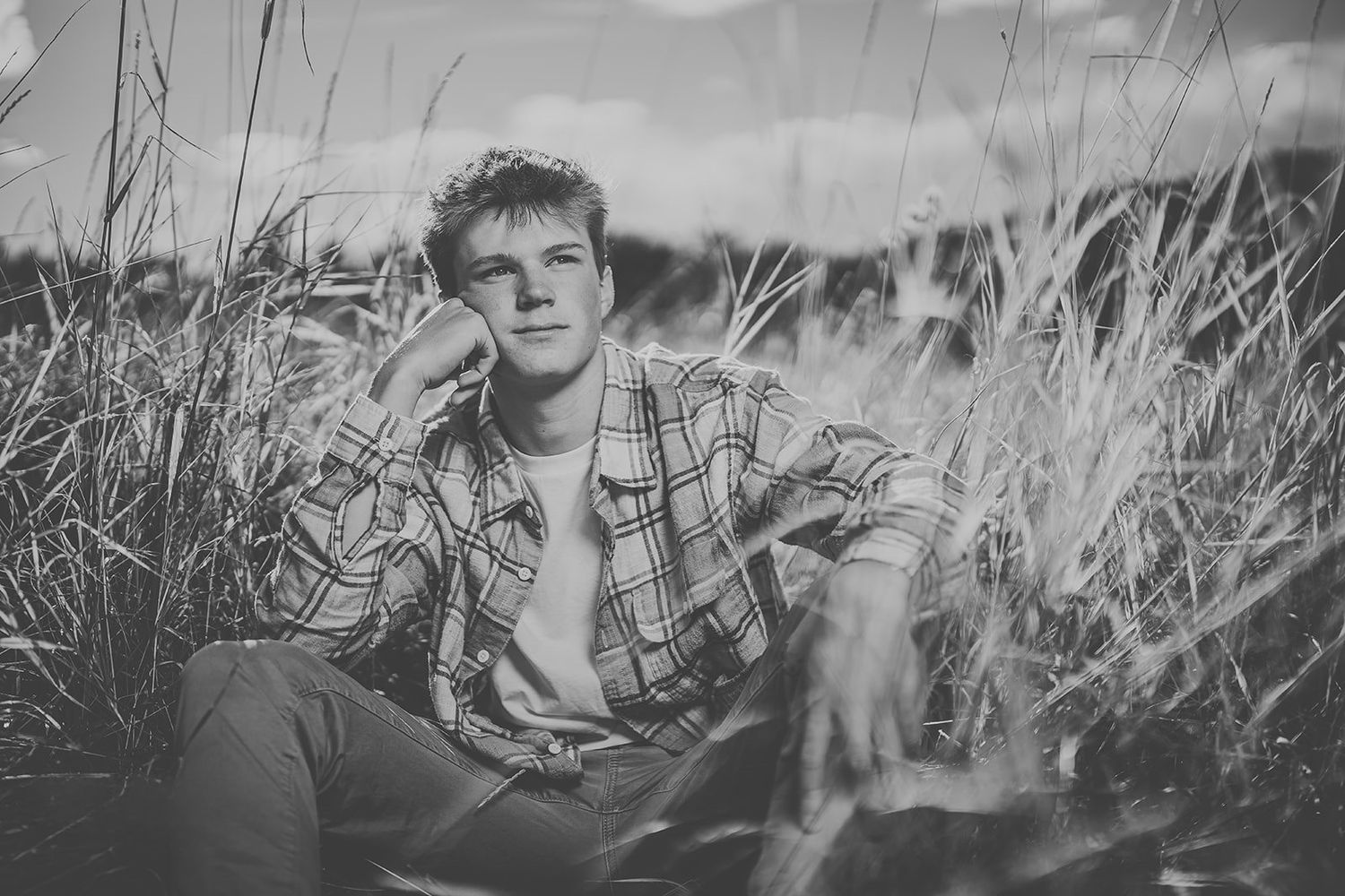 Young man in plaid shirt sitting relaxed in tall grass field, black and white portrait photography.