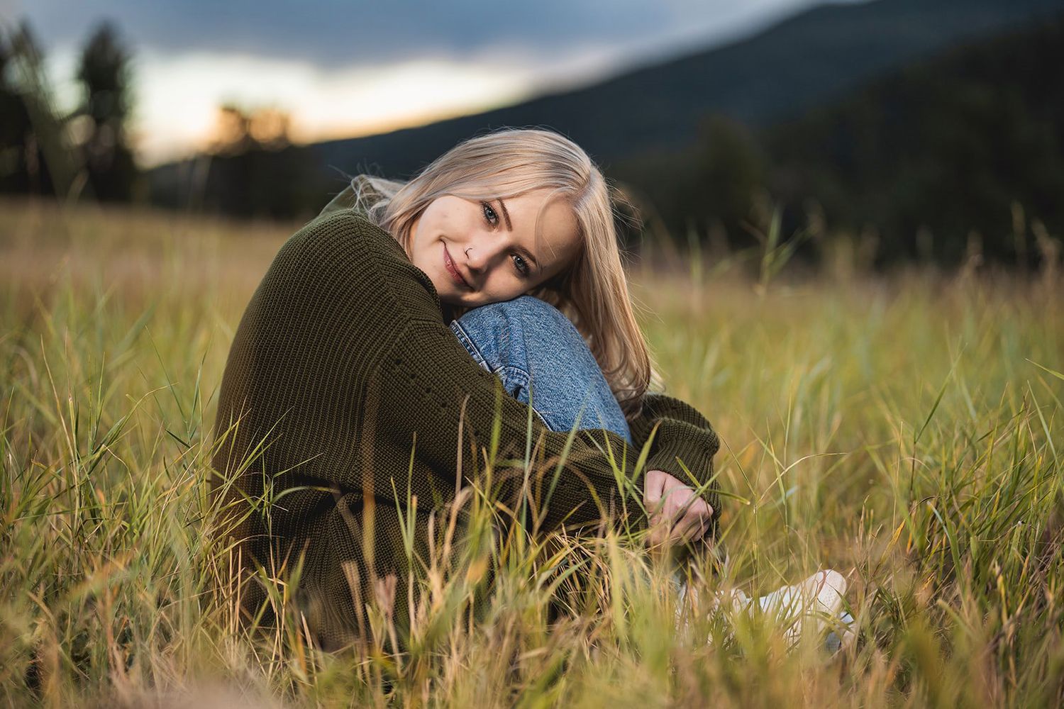 Blonde woman hugging her knees and smiling softly while sitting in a grassy autumn field.
