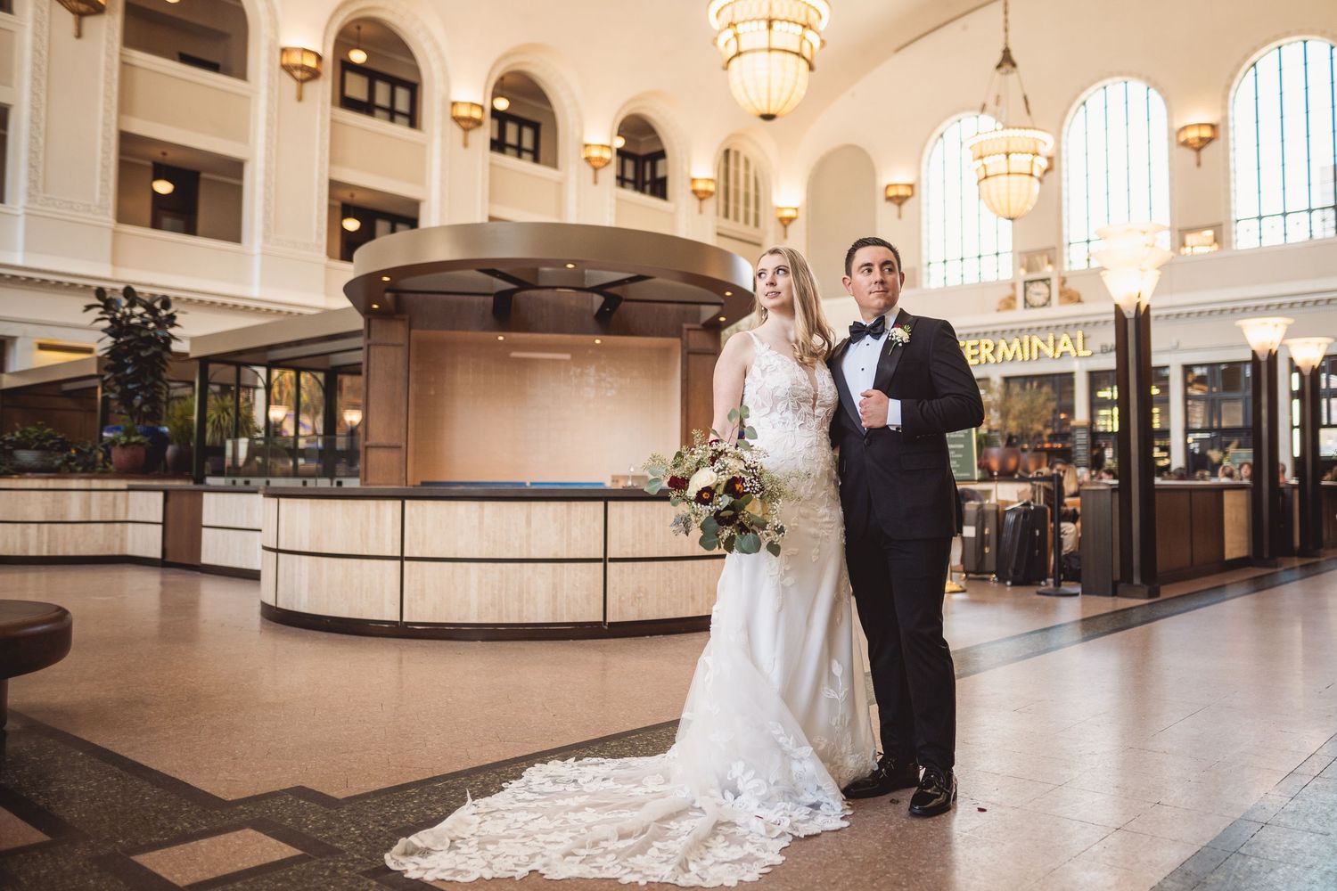 Bride and groom posing elegantly in a grand historic train terminal with ornate chandeliers on their wedding day.
