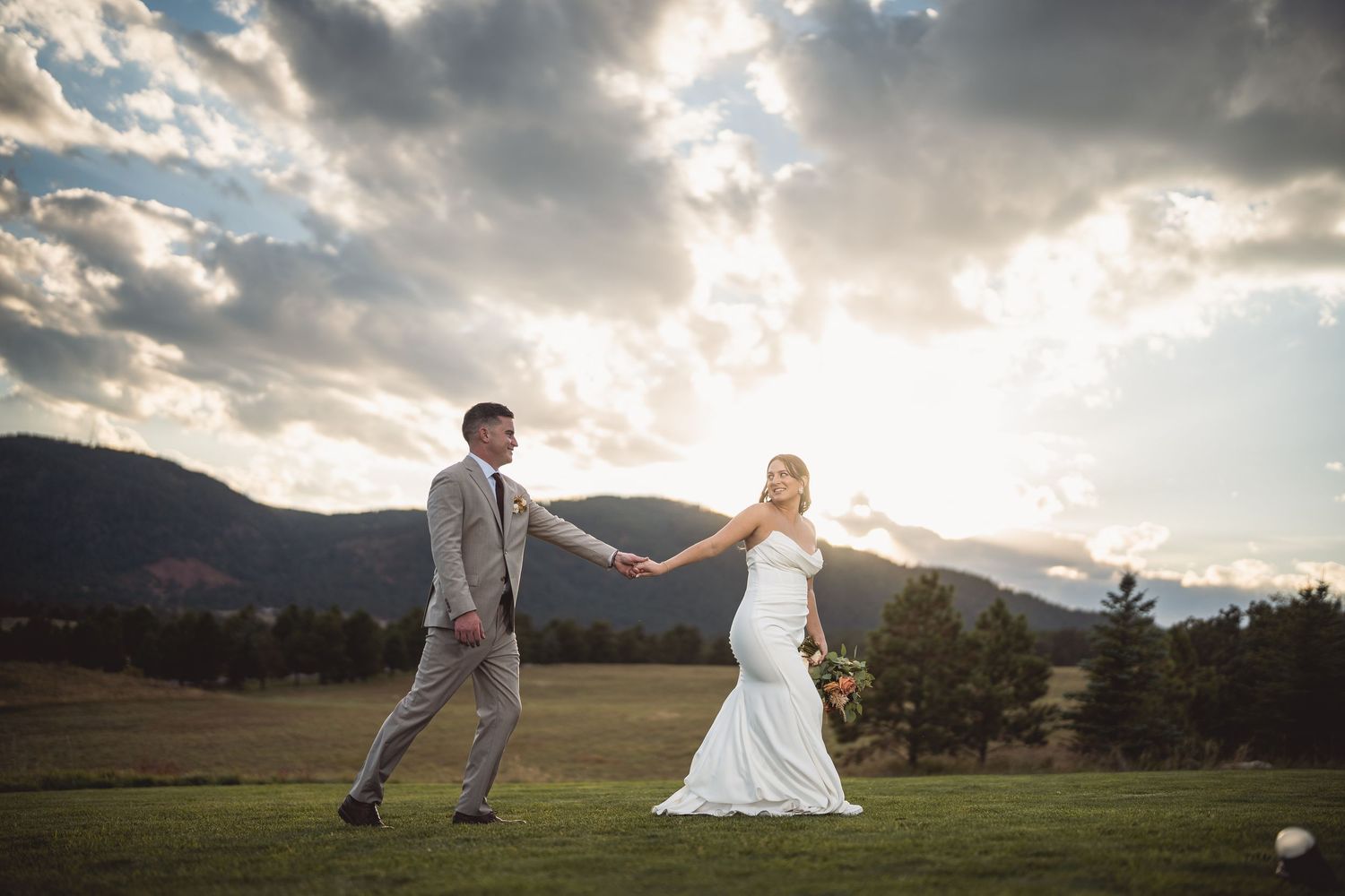 Bride and groom hold hands at sunset on a golf course with mountain backdrop on their wedding day.