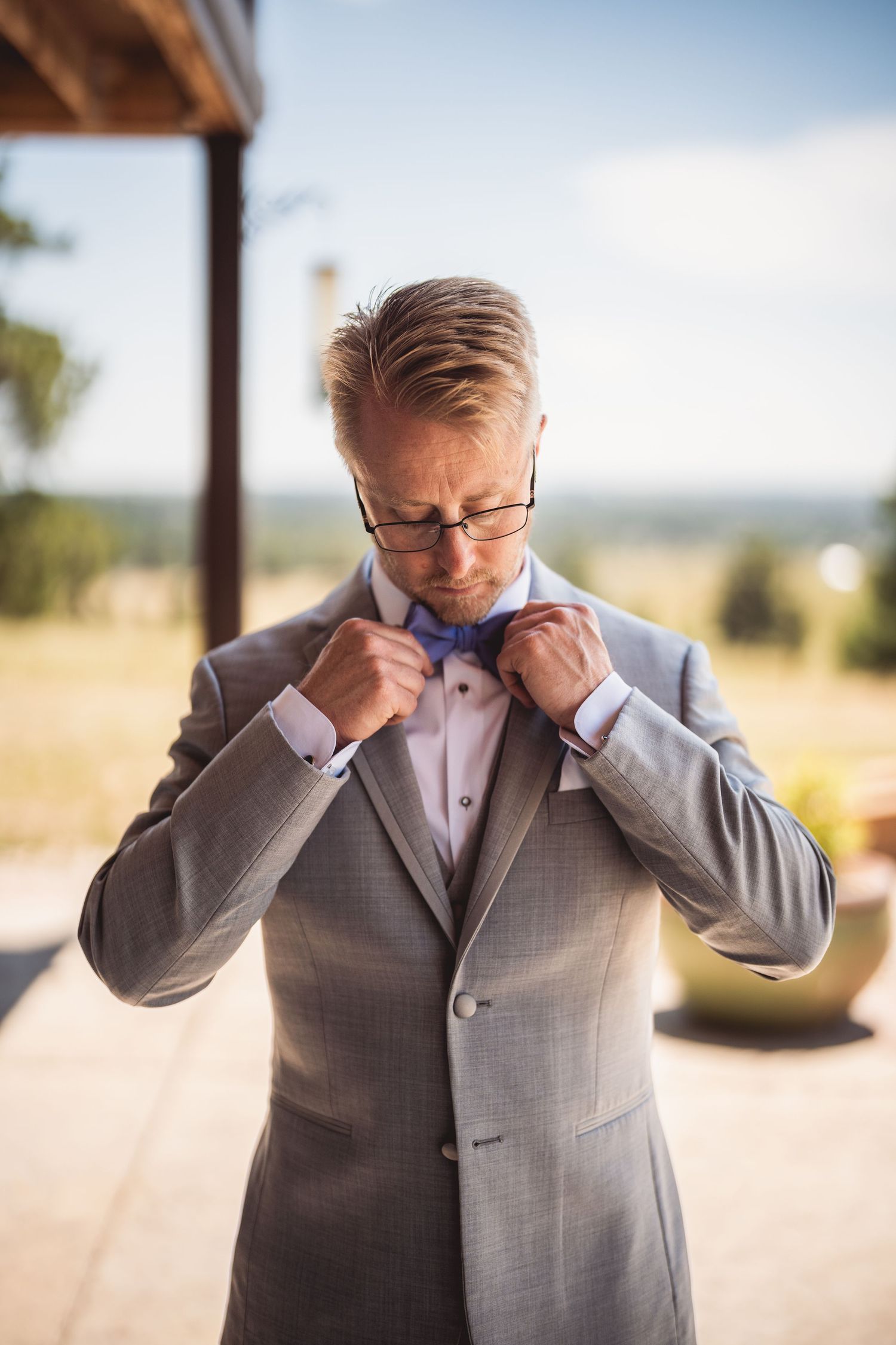 Groom in gray suit adjusting blue bow tie outdoors on sunny day.