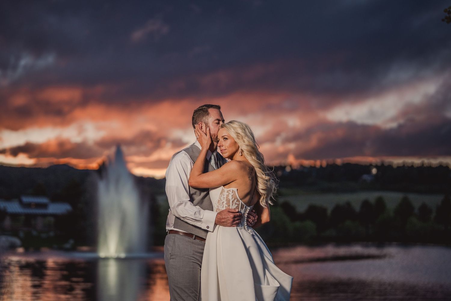 Couple embracing at sunset near a fountain, dramatic purple and orange sky, wedding portrait photography.