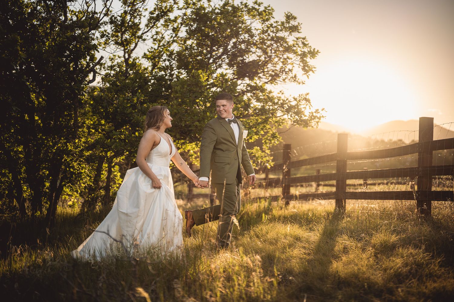 Bride and groom hold hands walking through golden sunlit field beside rustic wooden fence on wedding day.