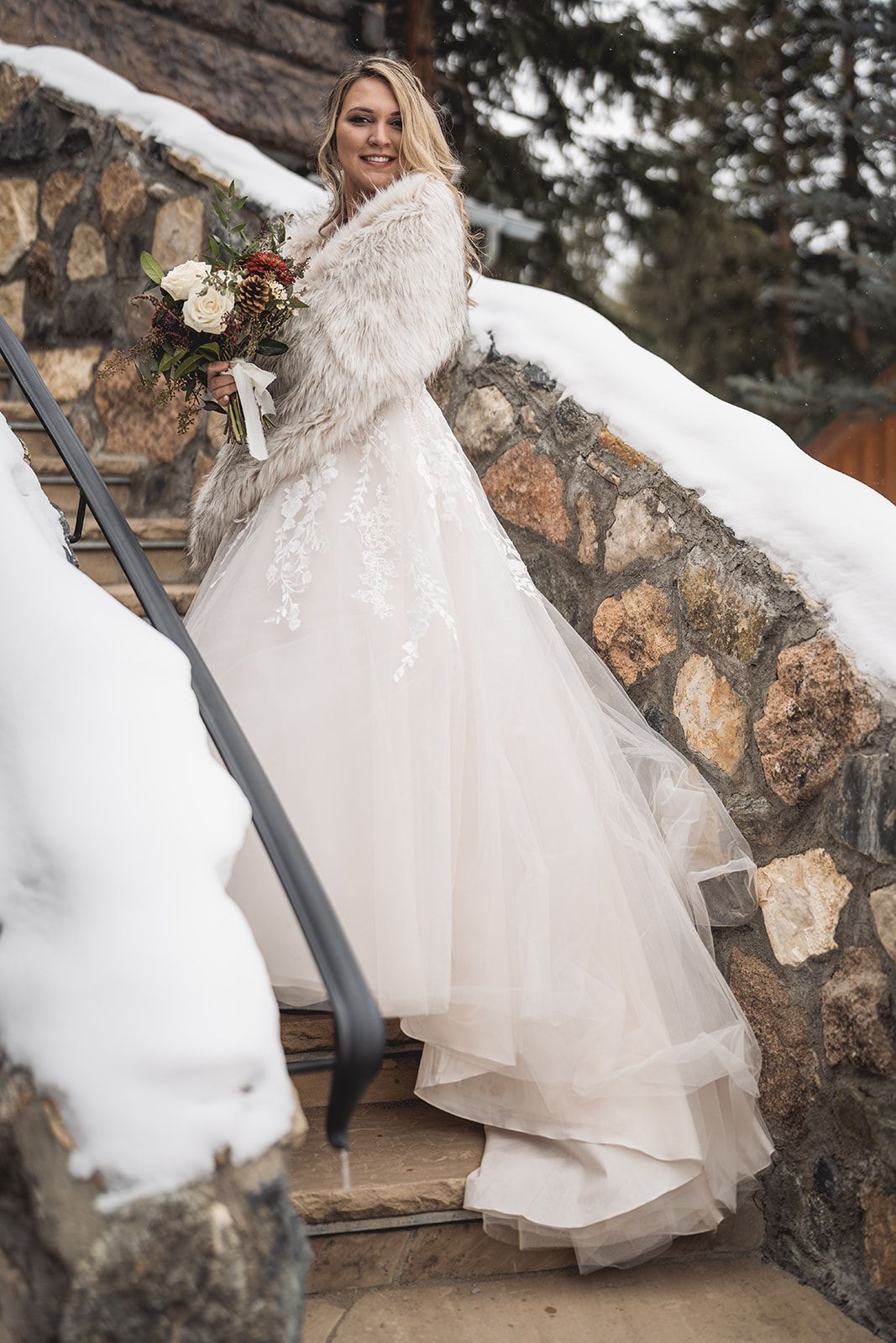 Bride in white lace gown and fur stole holds bouquet on snowy stone steps in winter wedding setting.